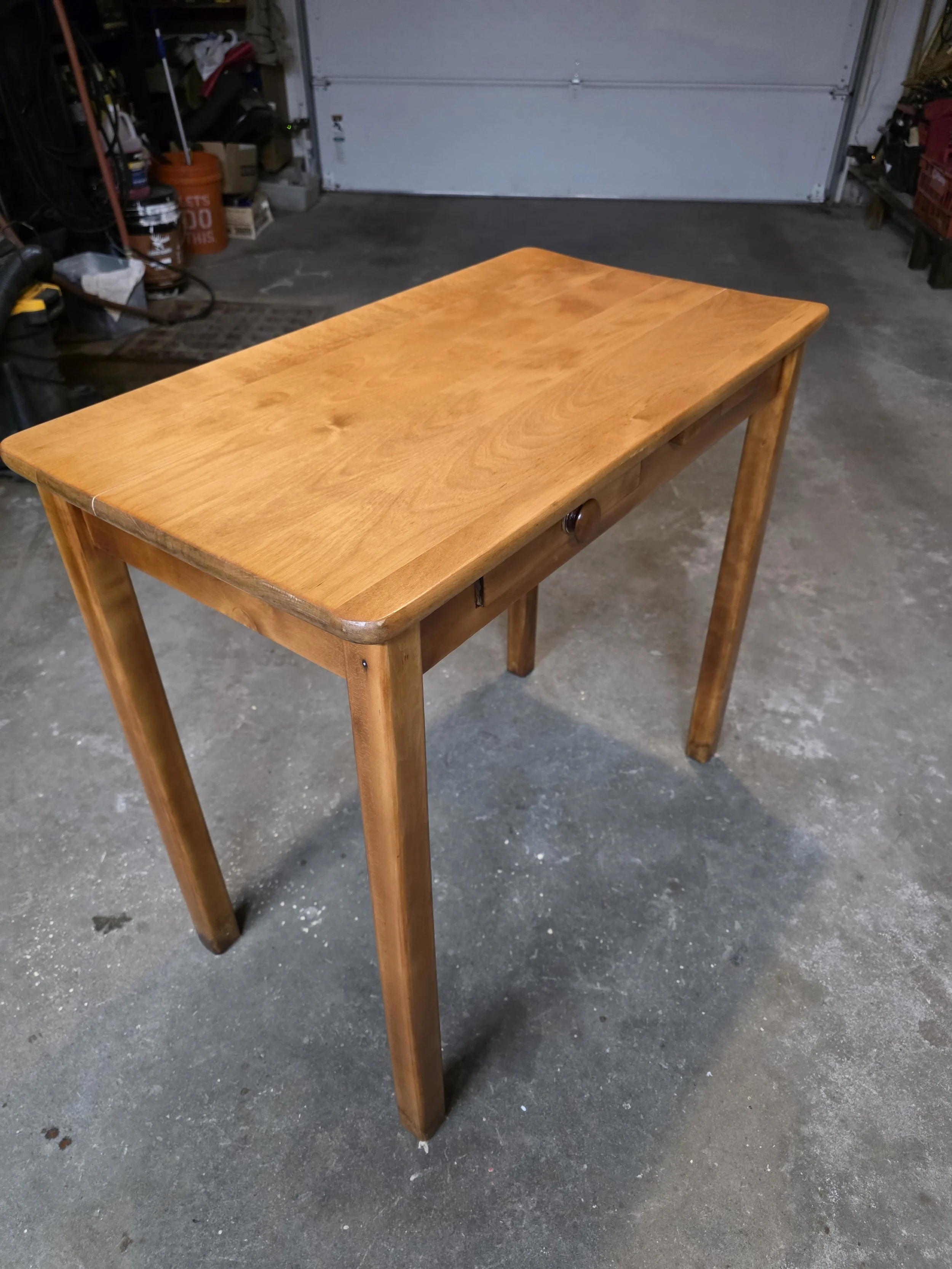 A wooden table with four legs, situated in a garage with a concrete floor and various items stored on shelves in the background.