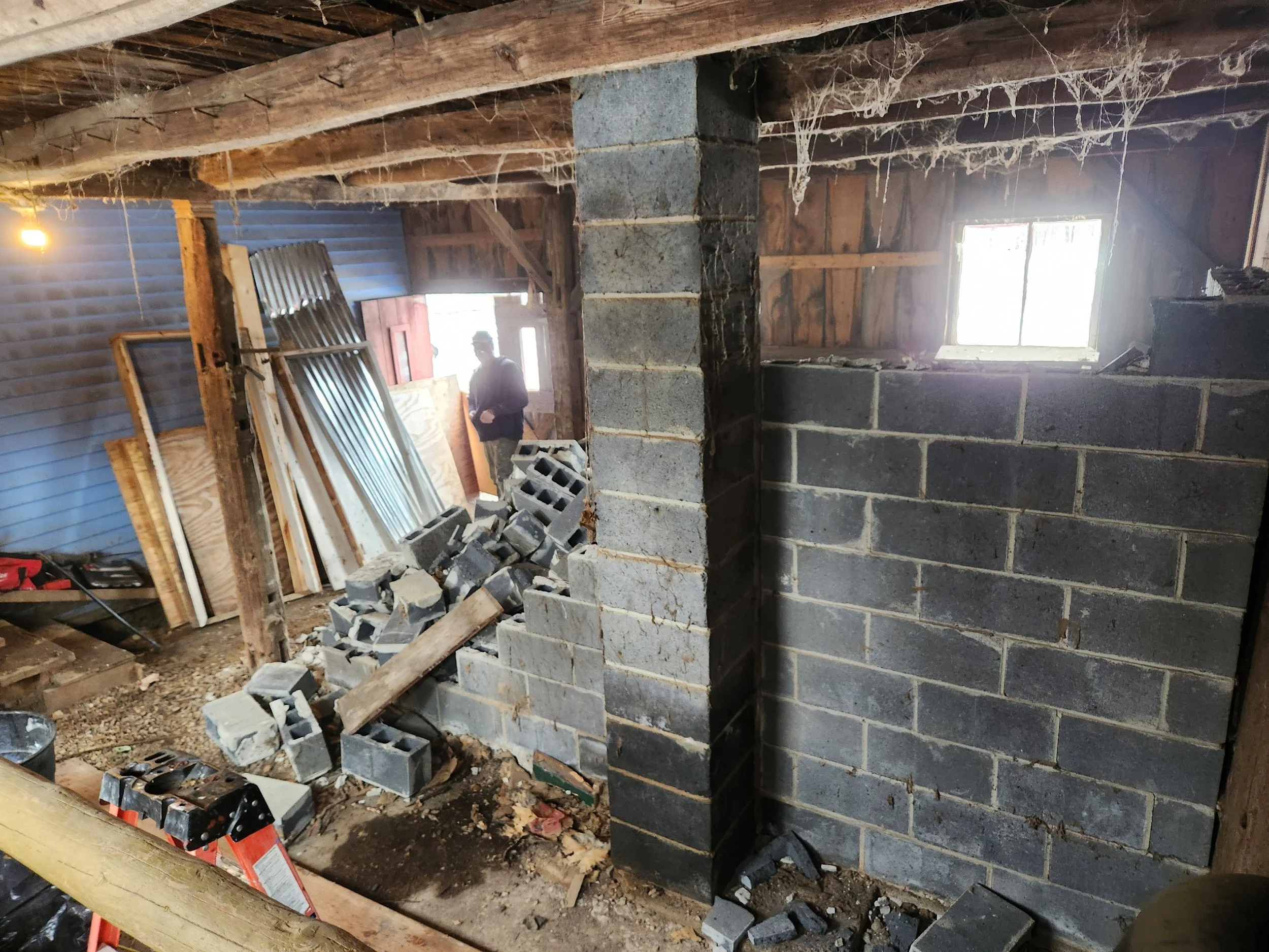 Interior of a construction site with cinder block walls, a pile of bricks, wooden beams, a person working in the background, and tools on the ground.