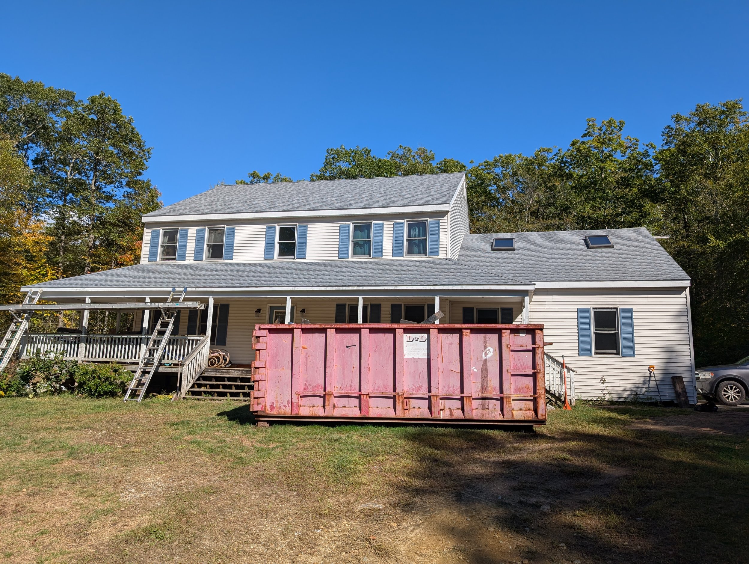 A large house with a two-story section, white siding, and blue shutters. There is a wooden deck with a staircase, a pink construction dumpster in the front yard, and a car parked to the right. The background features trees and a clear blue sky.
