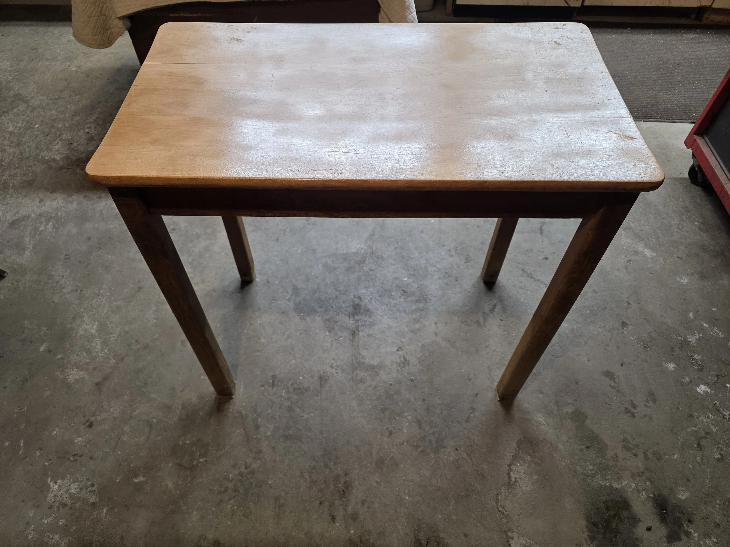 A wooden table with a light-colored top and darker legs, set on a concrete floor in a warehouse or workshop setting.