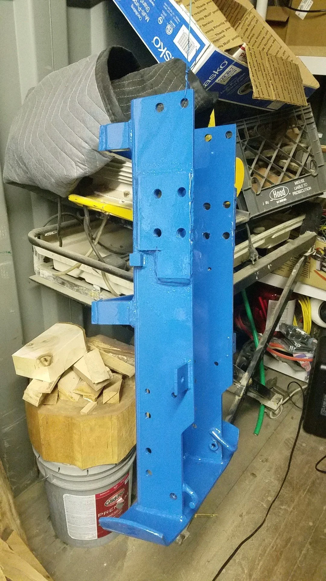 A large, blue-painted metal frame or bracket standing upright in a workshop or garage, with various tools, wood pieces, and supplies around it.