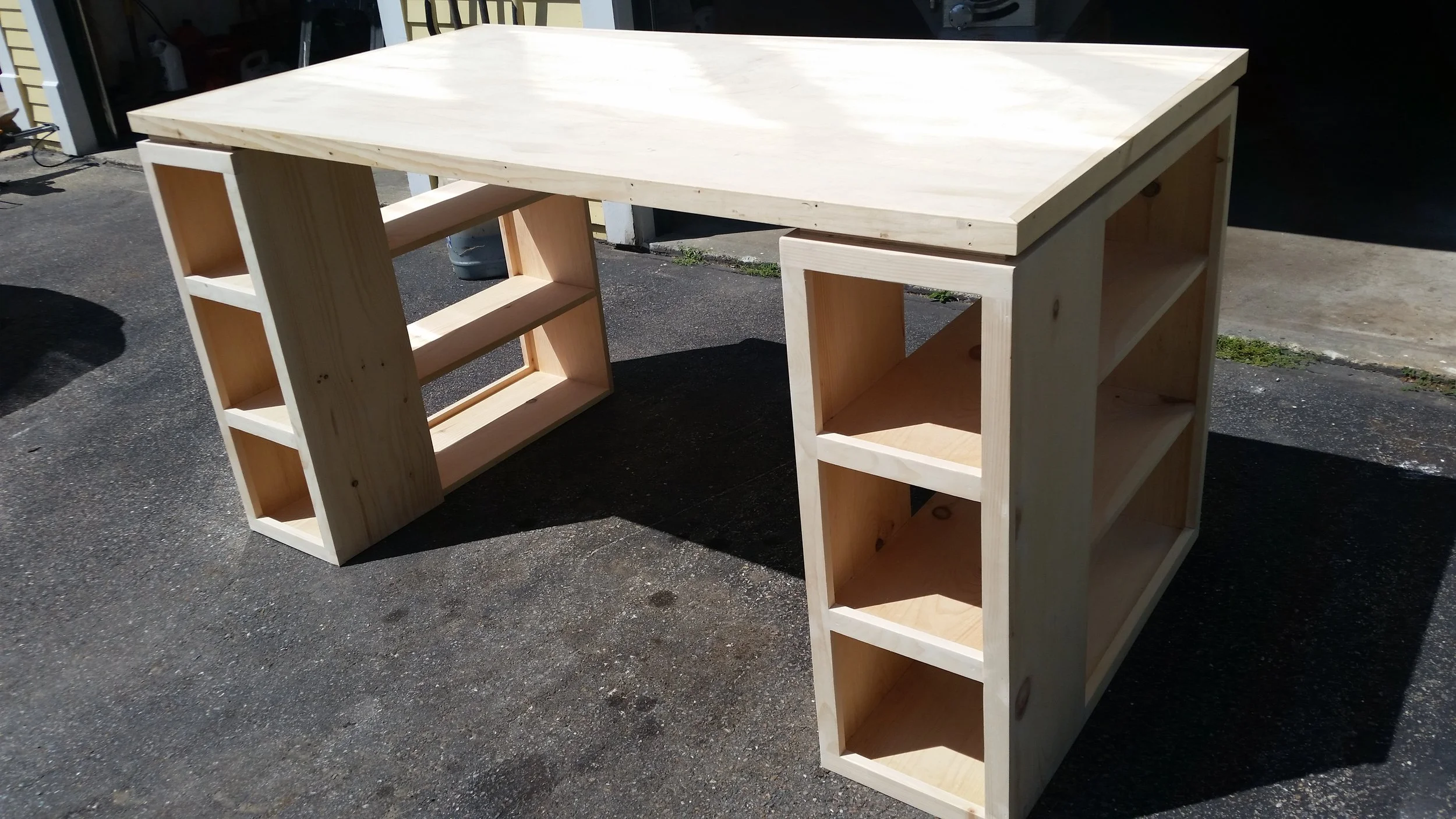 Unfinished wooden desk with open shelves on both sides, set outdoors on asphalt with sunlight.