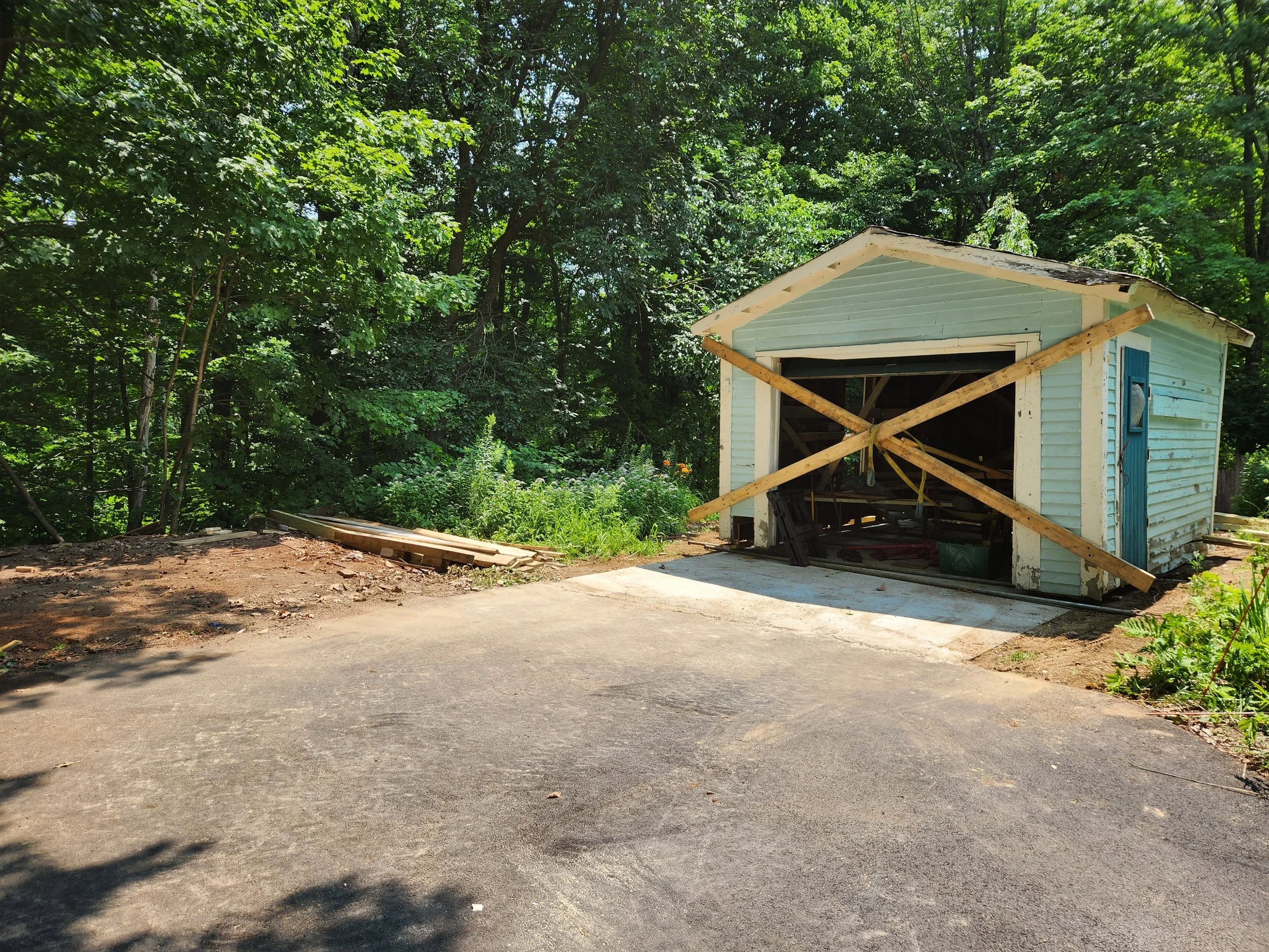 Garage under construction with wooden braces across the opening, in a wooded area with a paved driveway.