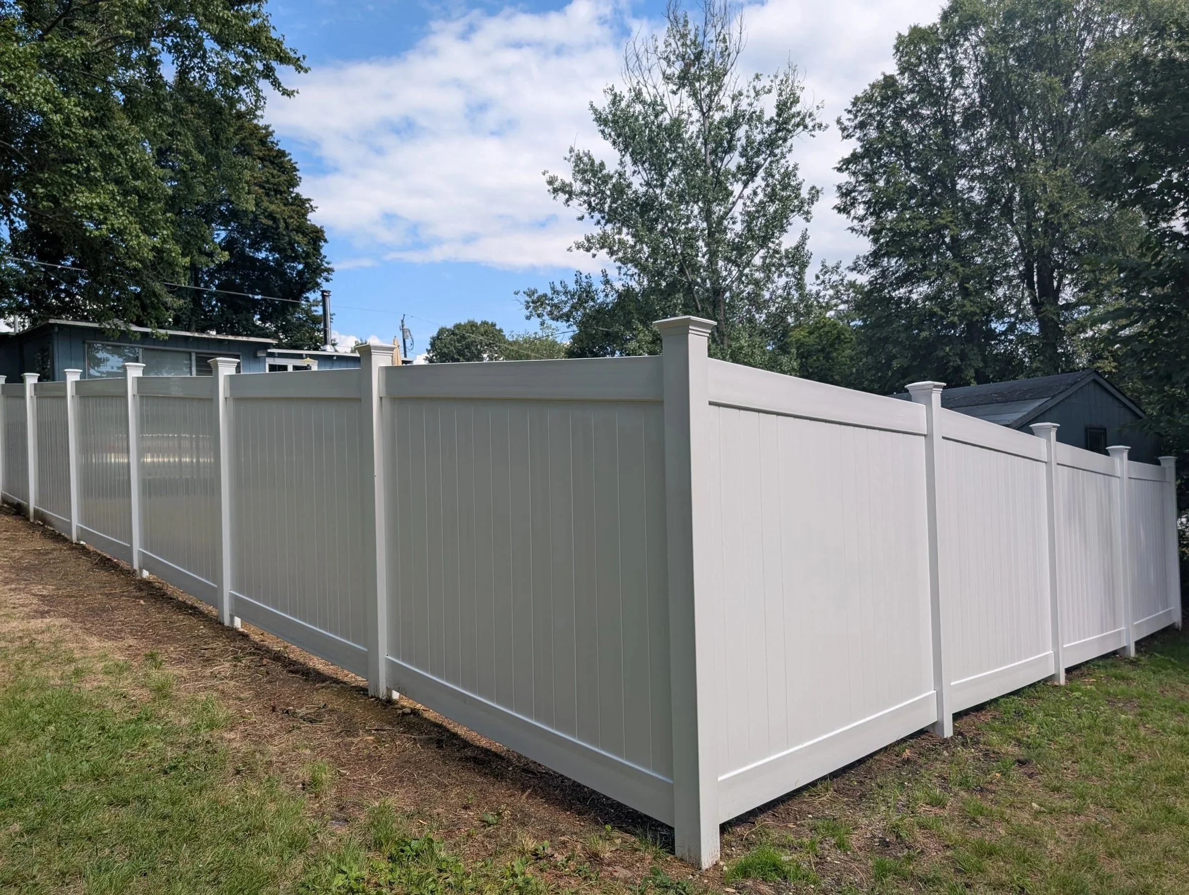 White vinyl privacy fence enclosing a backyard area, with trees and a house in the background under a partly cloudy sky.