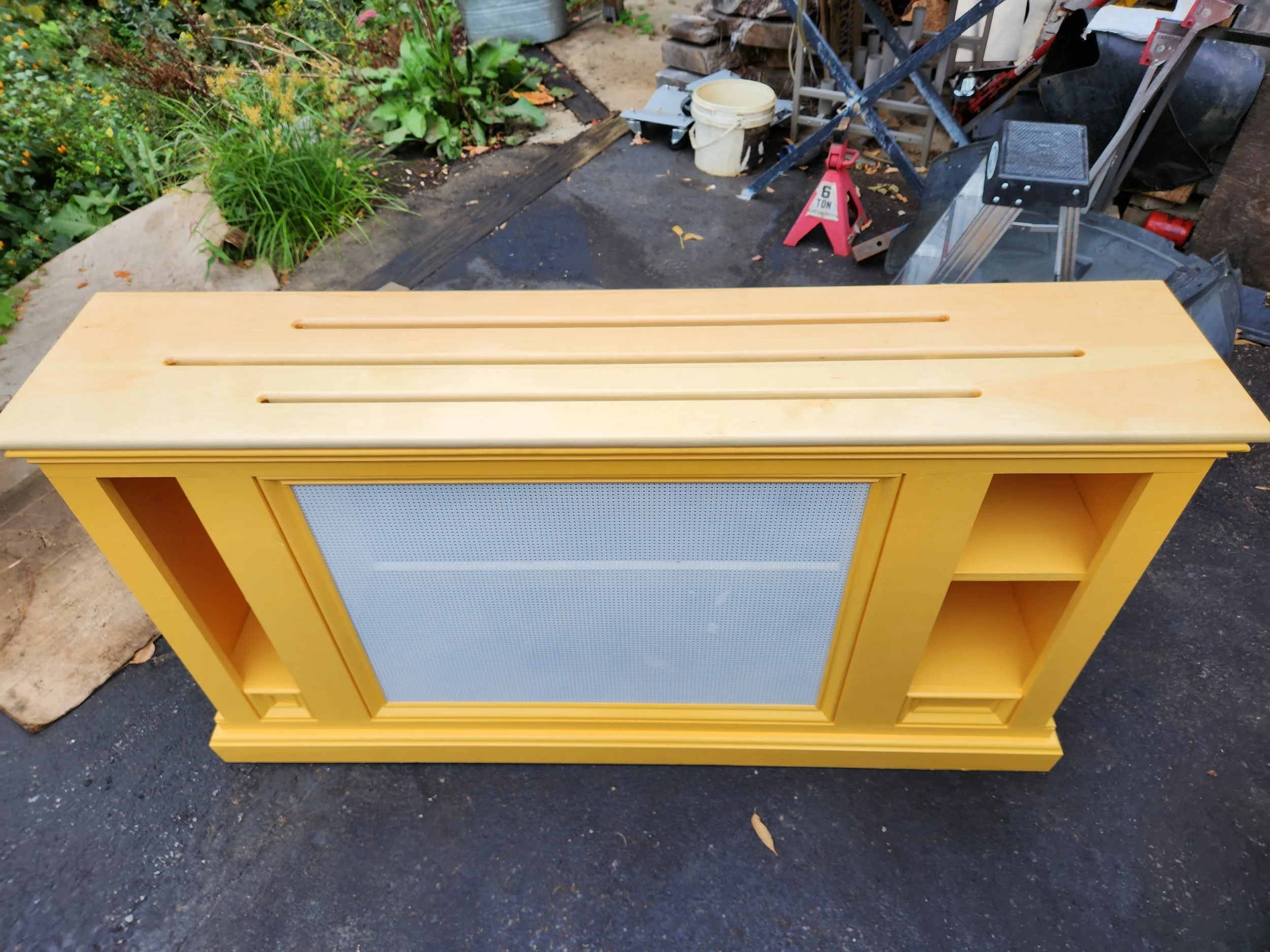 Yellow-painted wooden cabinet with a sliding glass door in the center, featuring three open shelves on the right side and a small compartment on the left side, placed outdoors on pavement with gardening tools and plants in the background.