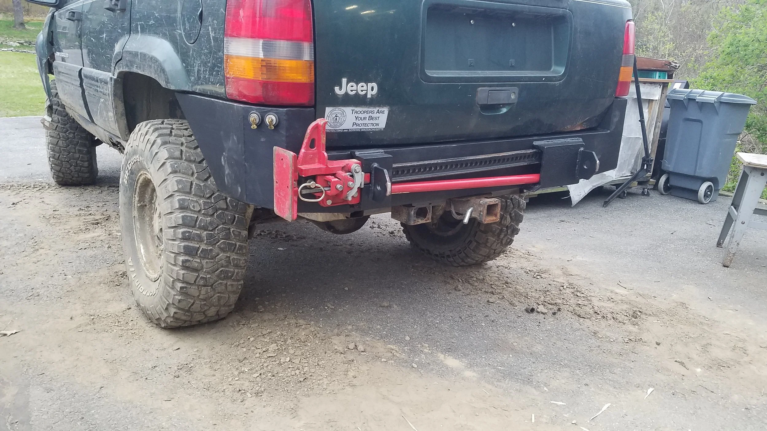 Back of a Jeep with off-road tires, a red recovery hook, and a hitch. There are trash bins and a small table nearby.