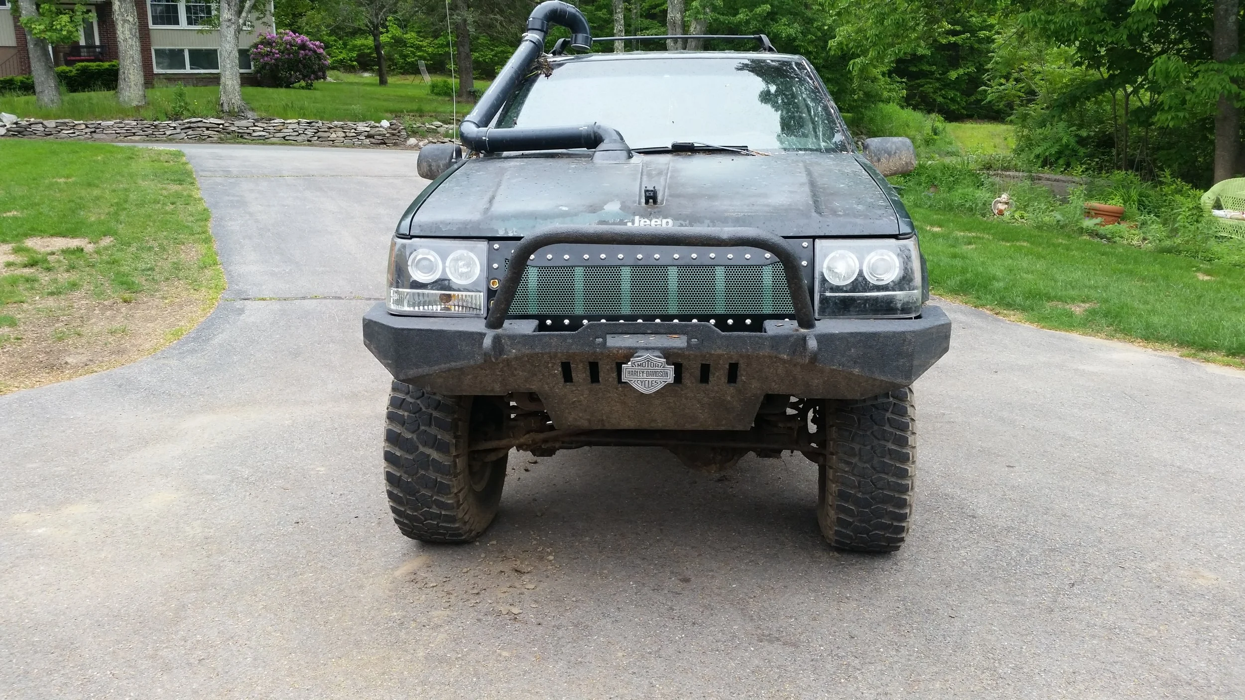 Front view of a black modified off-road Jeep with large tires, aftermarket bumper, grille guard, and snorkel, parked on a driveway surrounded by greenery.