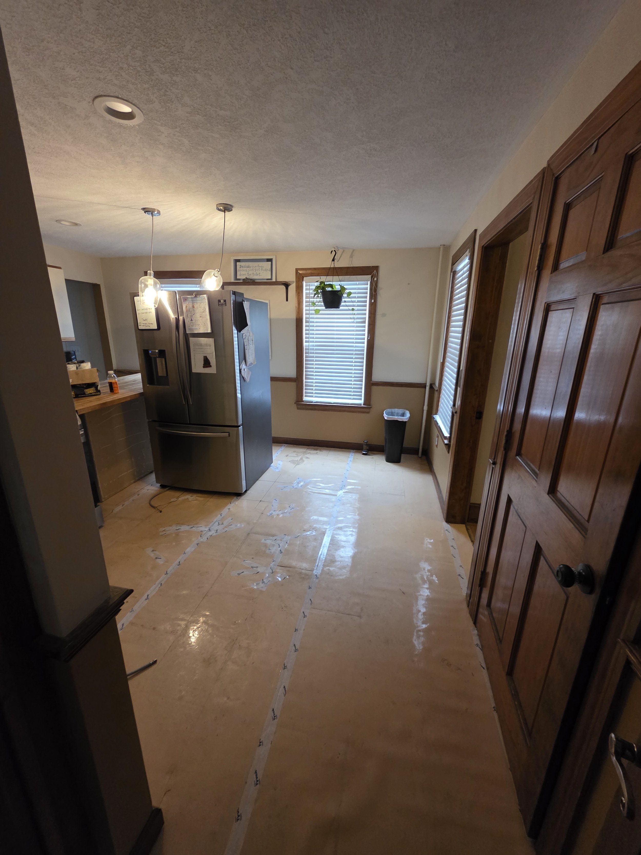 Empty kitchen with a plywood floor, refrigerator, and windows with blinds, during renovation.