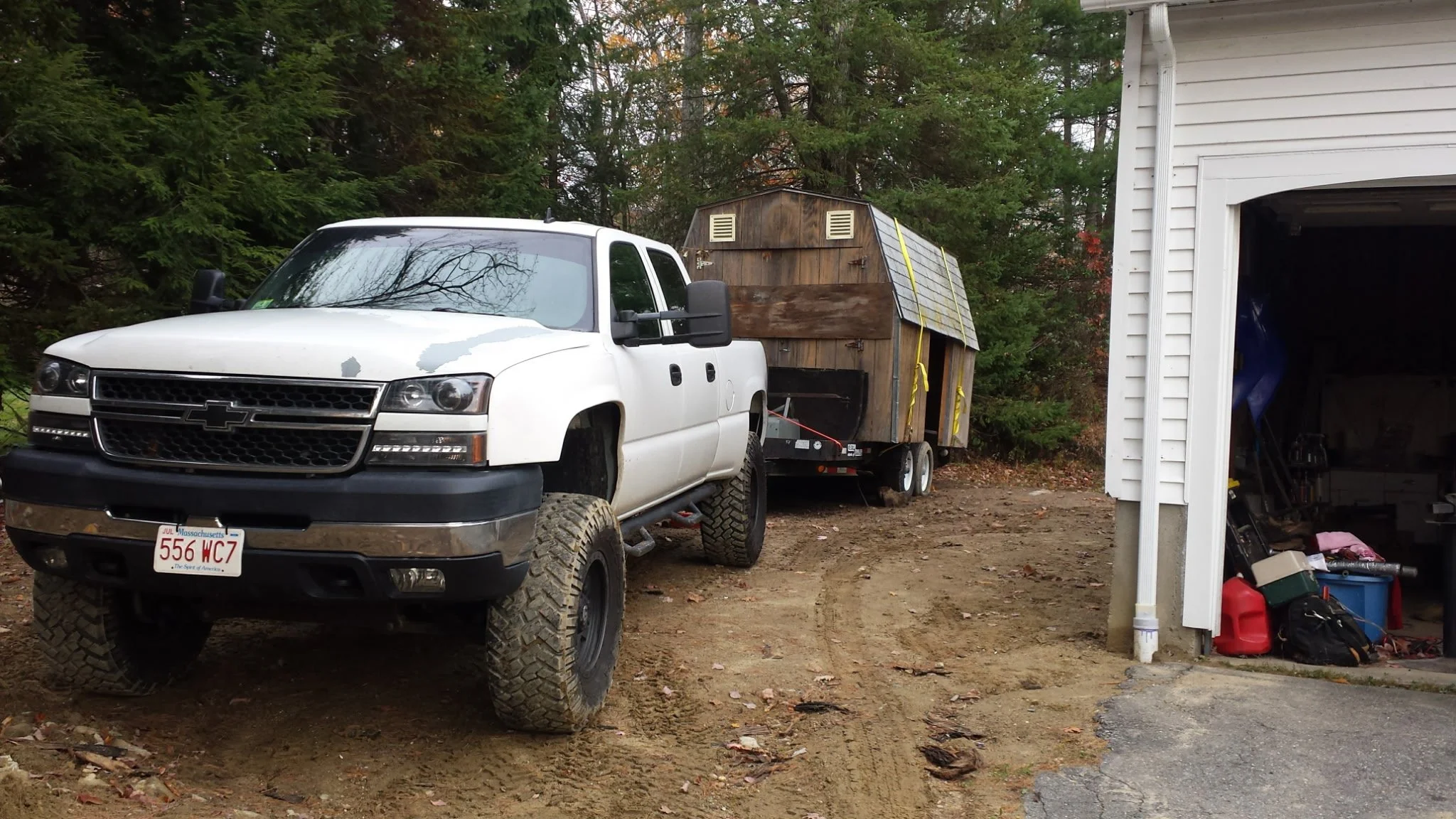 White Chevrolet pickup truck with off-road tires, parked near a garage, pulling a small wooden camper trailer on a dirt surface, with trees and part of a house visible.