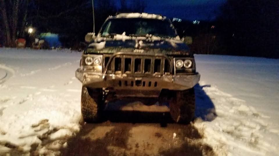 Front view of a black Jeep SUV with a damaged front bumper, parked on a snow-covered ground at night.