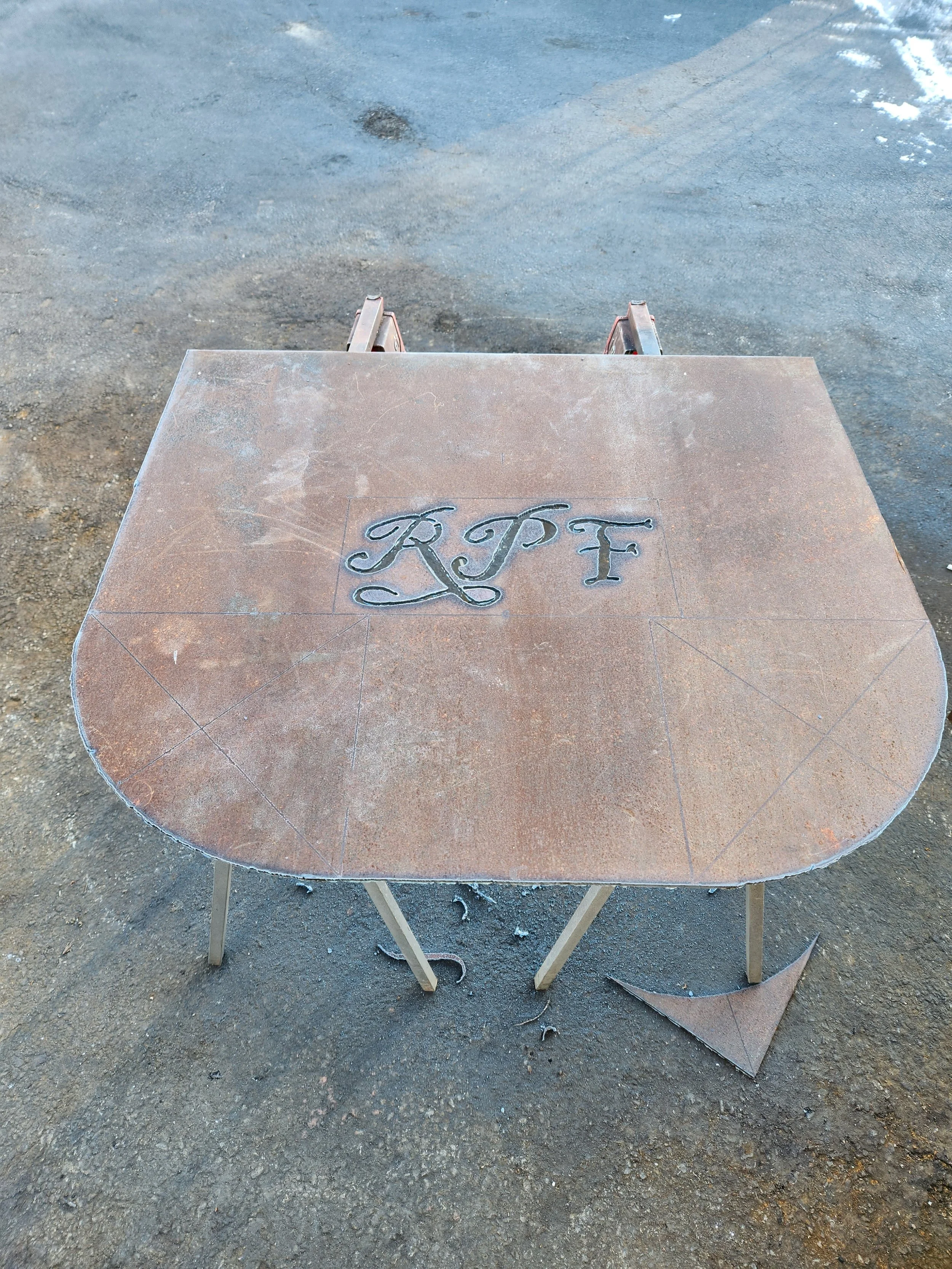 A folding table with a brownish surface displaying engraved initials 'RJF' on top, set on a dirt ground.