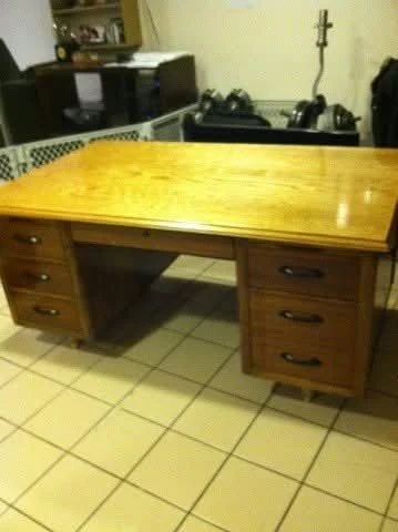 A wooden office desk with drawers on both sides, placed on a tiled floor in a room with kitchen appliances and other furniture in the background.