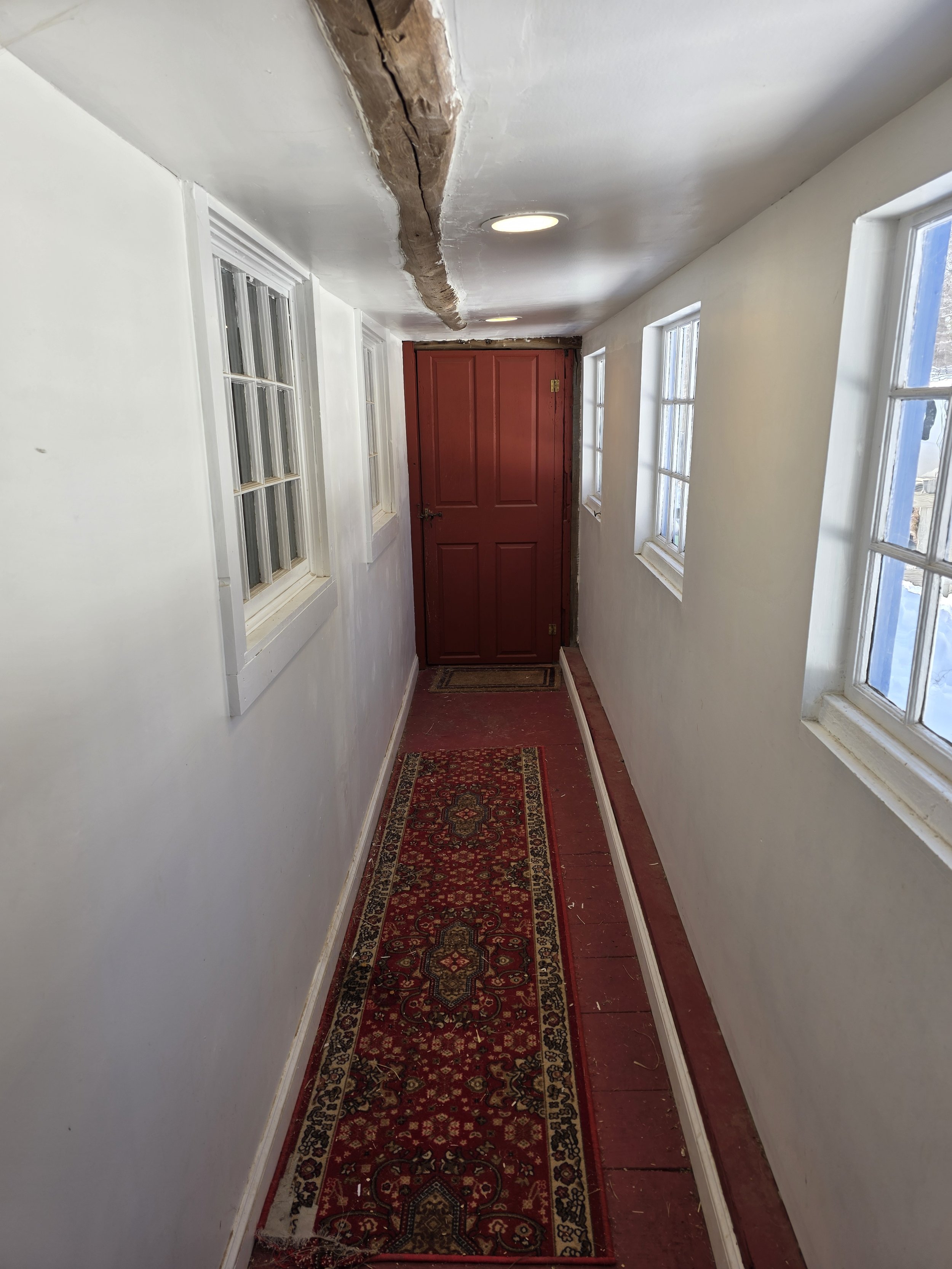 A narrow hallway with white walls, three double-hung windows on the right side, a red door at the end, a decorative red and black runner rug, and a partially damaged ceiling with exposed wooden beam.