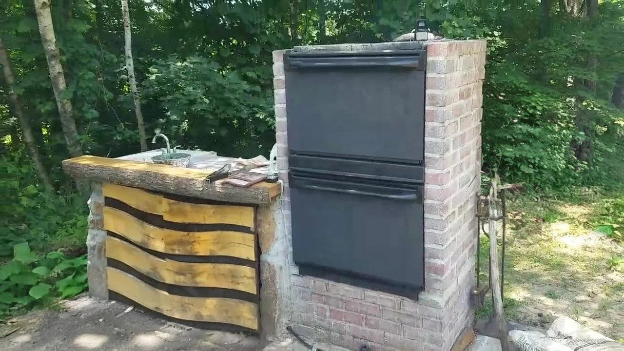 An outdoor outdoor kitchen featuring a brick oven, a rustic wooden countertop sink, and a faucet with trees and greenery in the background.