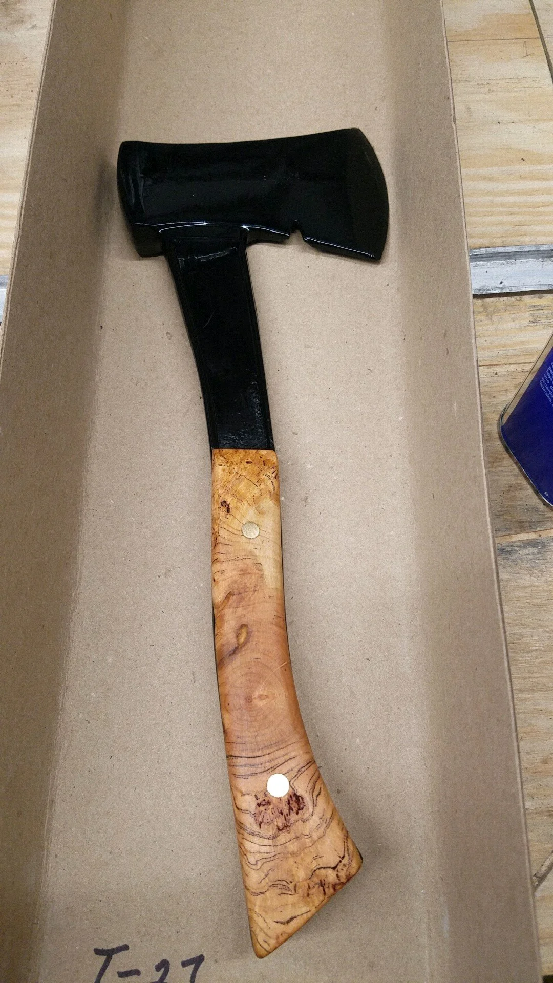 A hatchet with a black metal head and a wooden handle, placed inside a cardboard box.