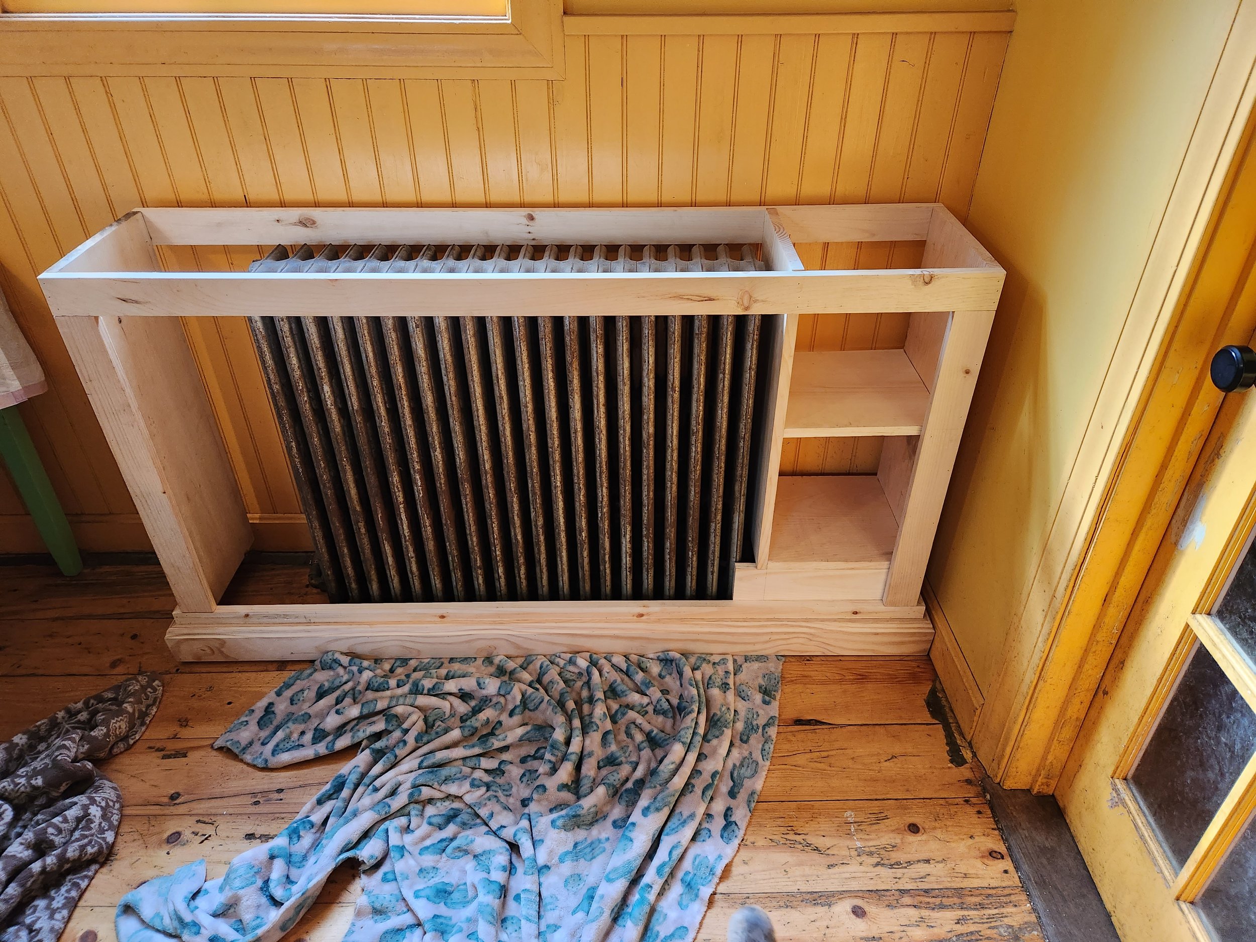 A partially assembled wooden radiator cover with open shelving on the right side, placed on a wooden floor in a room with yellow-painted walls and a glass door.