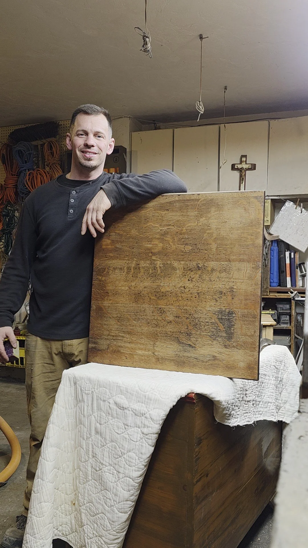 A man standing in a workshop with a wooden piece leaning against a piece of furniture covered with a quilted white cloth.