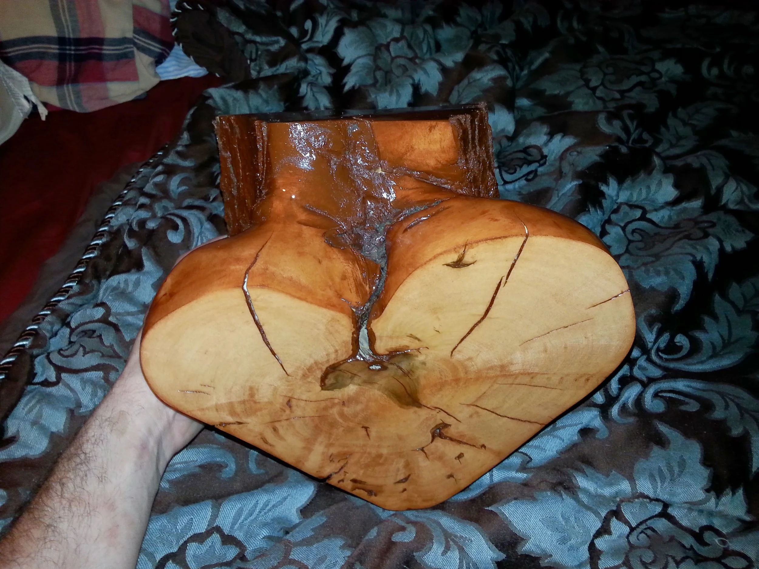 A wooden heart-shaped sculpture with a cracked, natural wood finish, held by a person's hand over a patterned fabric background.