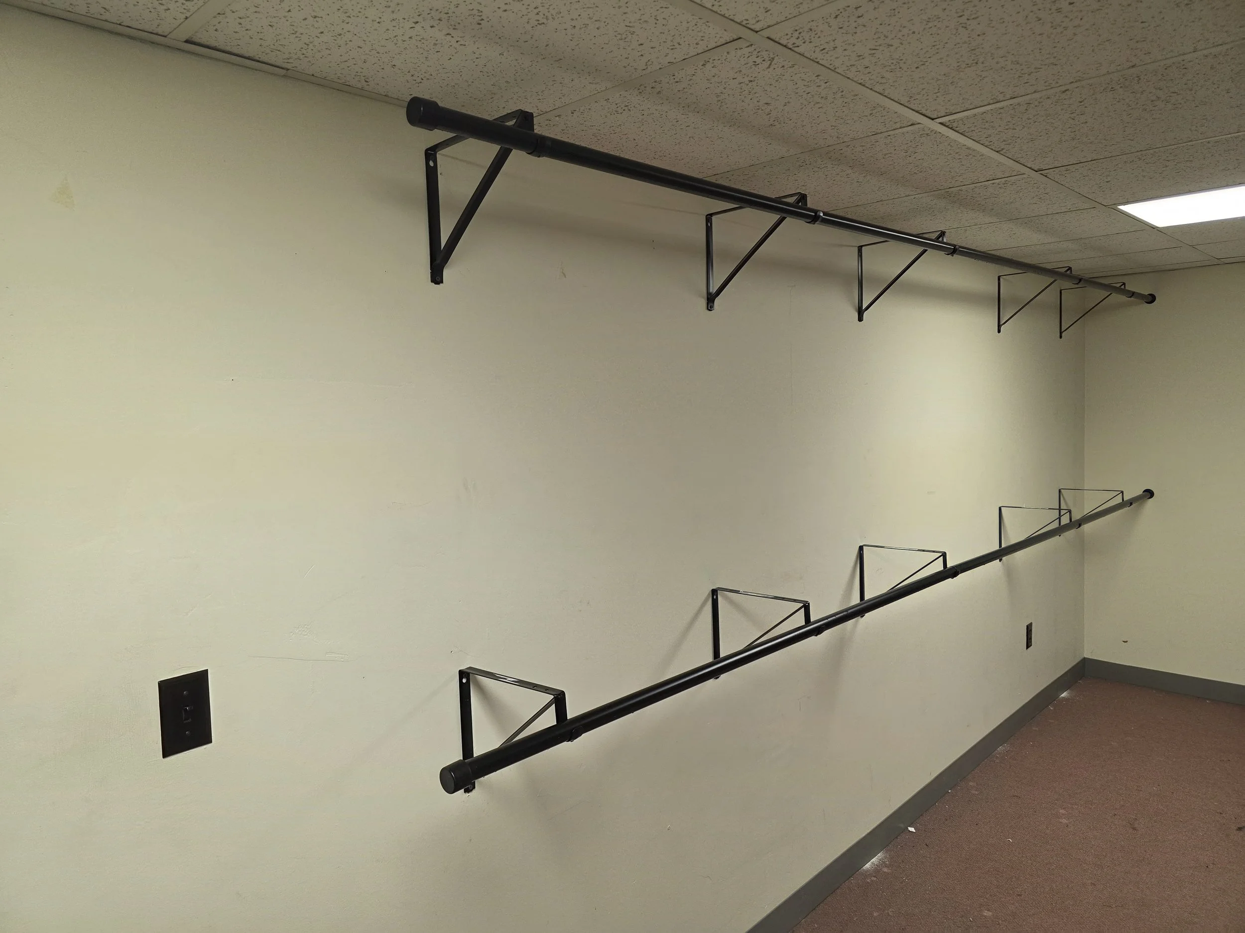 Empty room with beige walls, brown carpet, ceiling tiles, a black handrail on the wall, and a ceiling-mounted curtain rod with brackets.