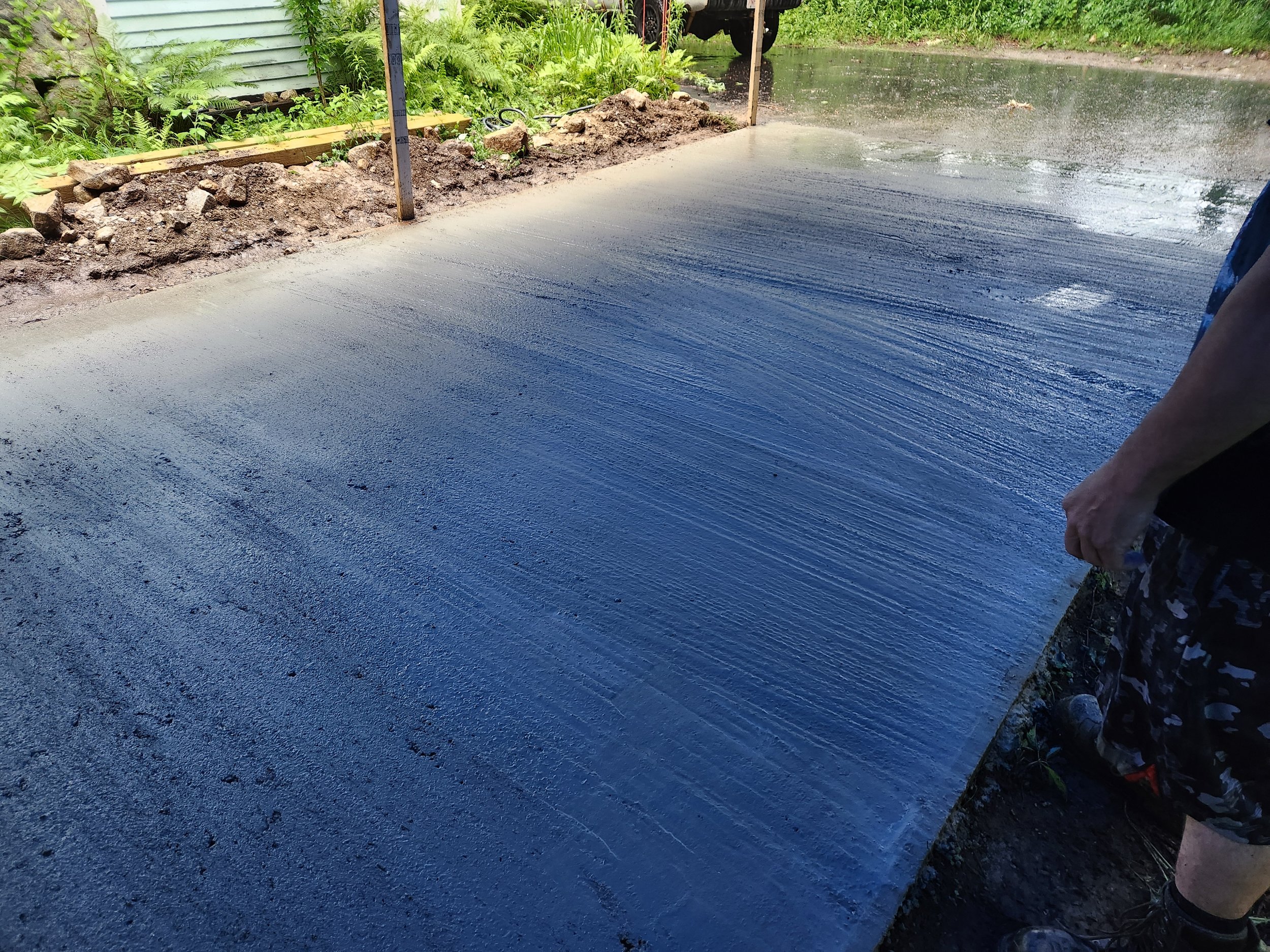 Person smoothing wet concrete in a freshly poured driveway or sidewalk with a trowel or float, surrounded by green plants and a rural setting.