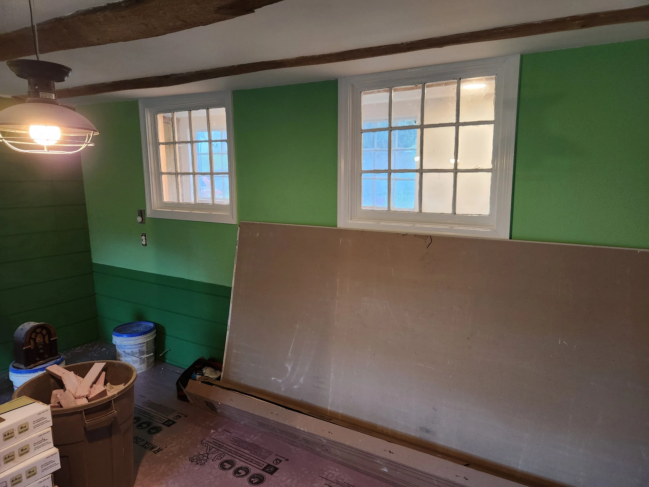 Room under renovation with green walls, two windows, construction materials, and a large board leaning against the wall.