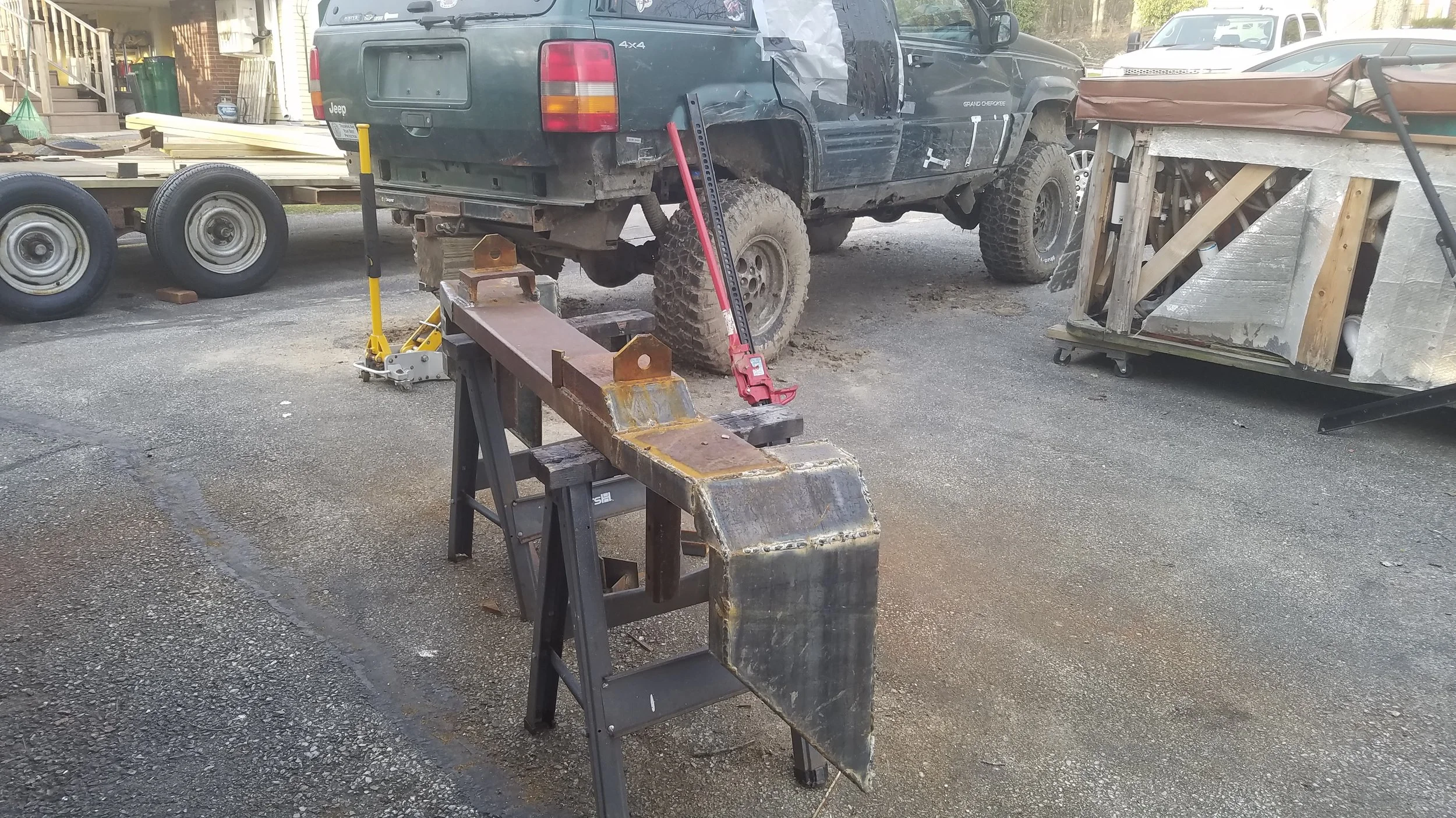 A workbench with metal pieces and tools in front of a black Jeep Grand Cherokee that is lifted on blocks, surrounded by various other vehicles and construction materials in a driveway or yard.