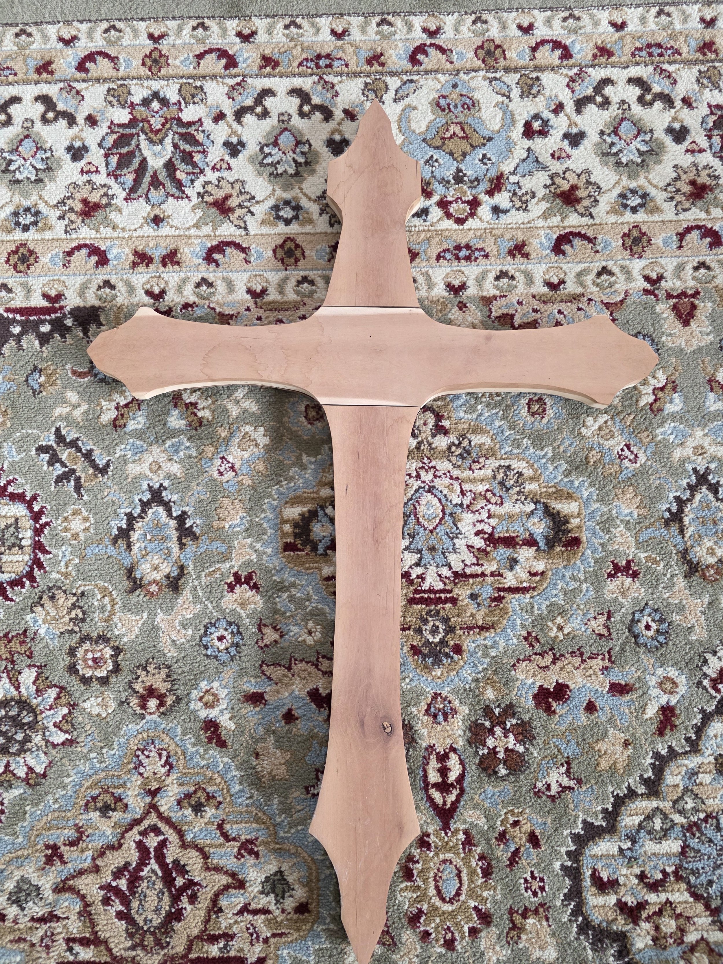 A wooden cross with decorative, pointed ends laying on a patterned rug with floral and geometric designs in beige, red, blue, and brown colors.