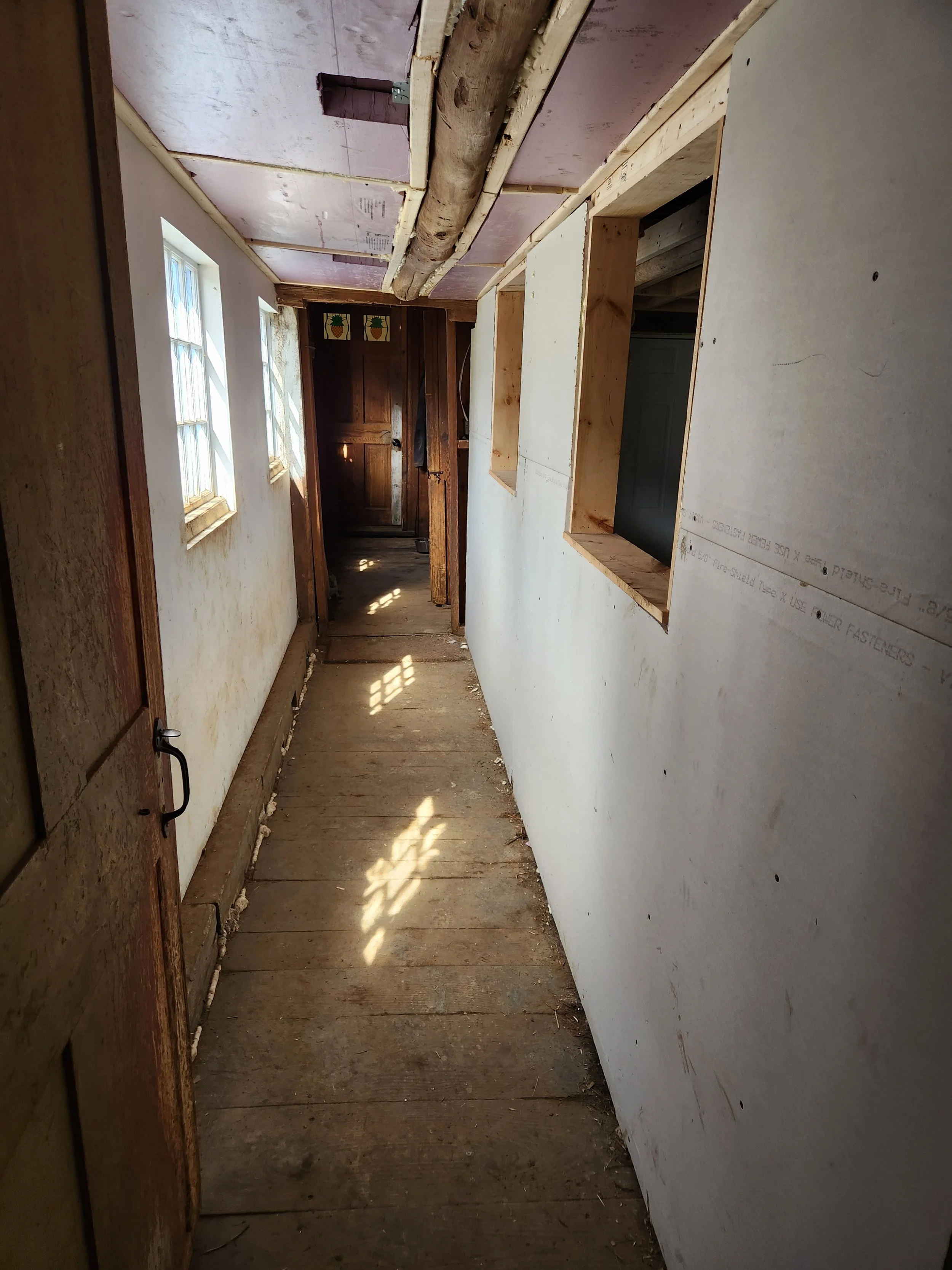 A narrow indoor hallway under renovation, with exposed wooden beams on the ceiling, unfinished walls, and a wooden floor. Sunlight streams through windows on the left, illuminating the space.