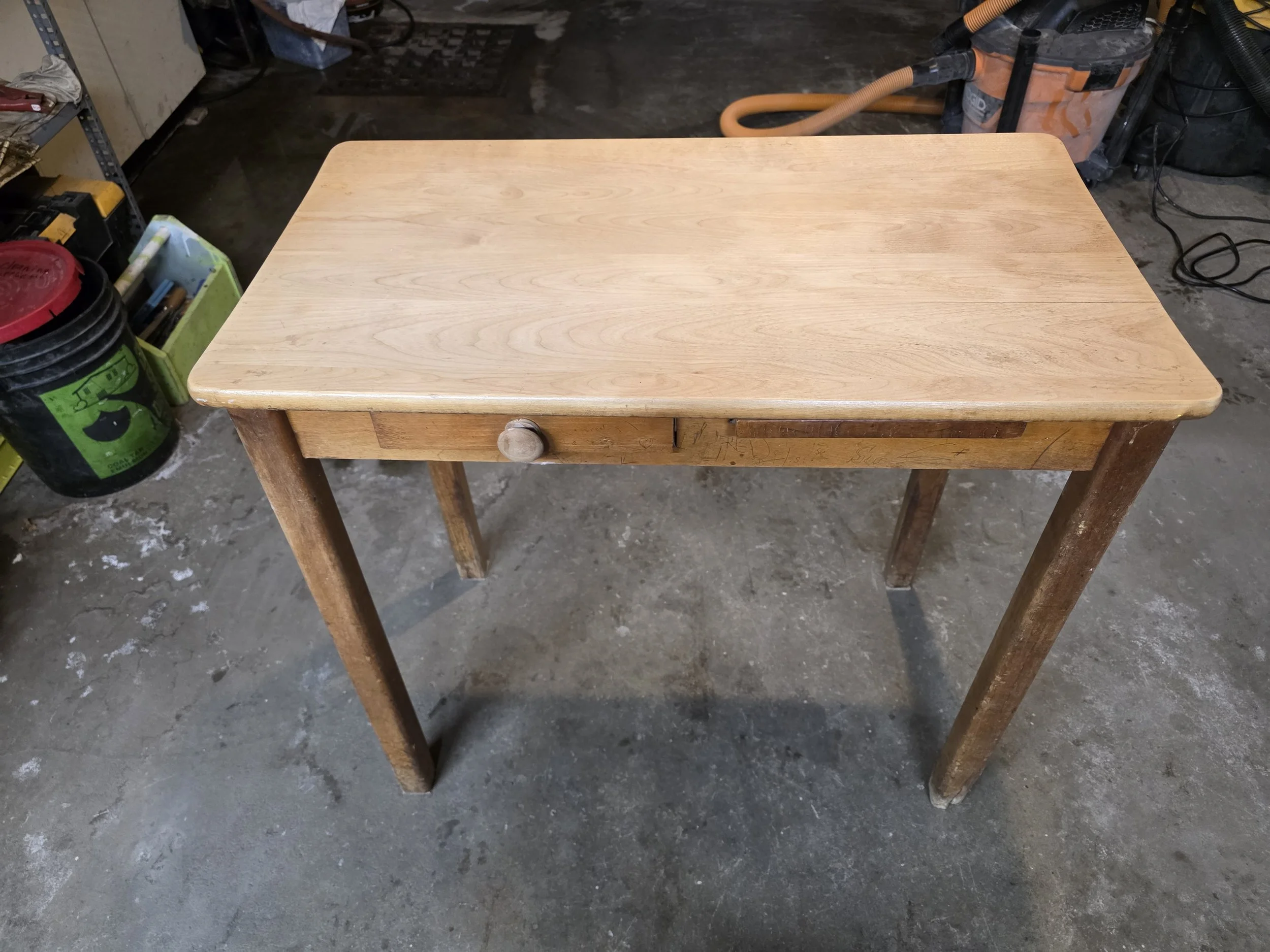 A small wooden table with a single drawer and turned wooden knob, placed on a concrete floor in a workshop or garage setting.