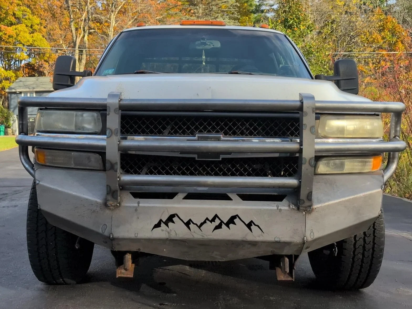 Front view of a silver Chevrolet police or rescue vehicle with a metal grille guard and a mountain decal on the lower bumper, parked on asphalt with autumn trees in the background.