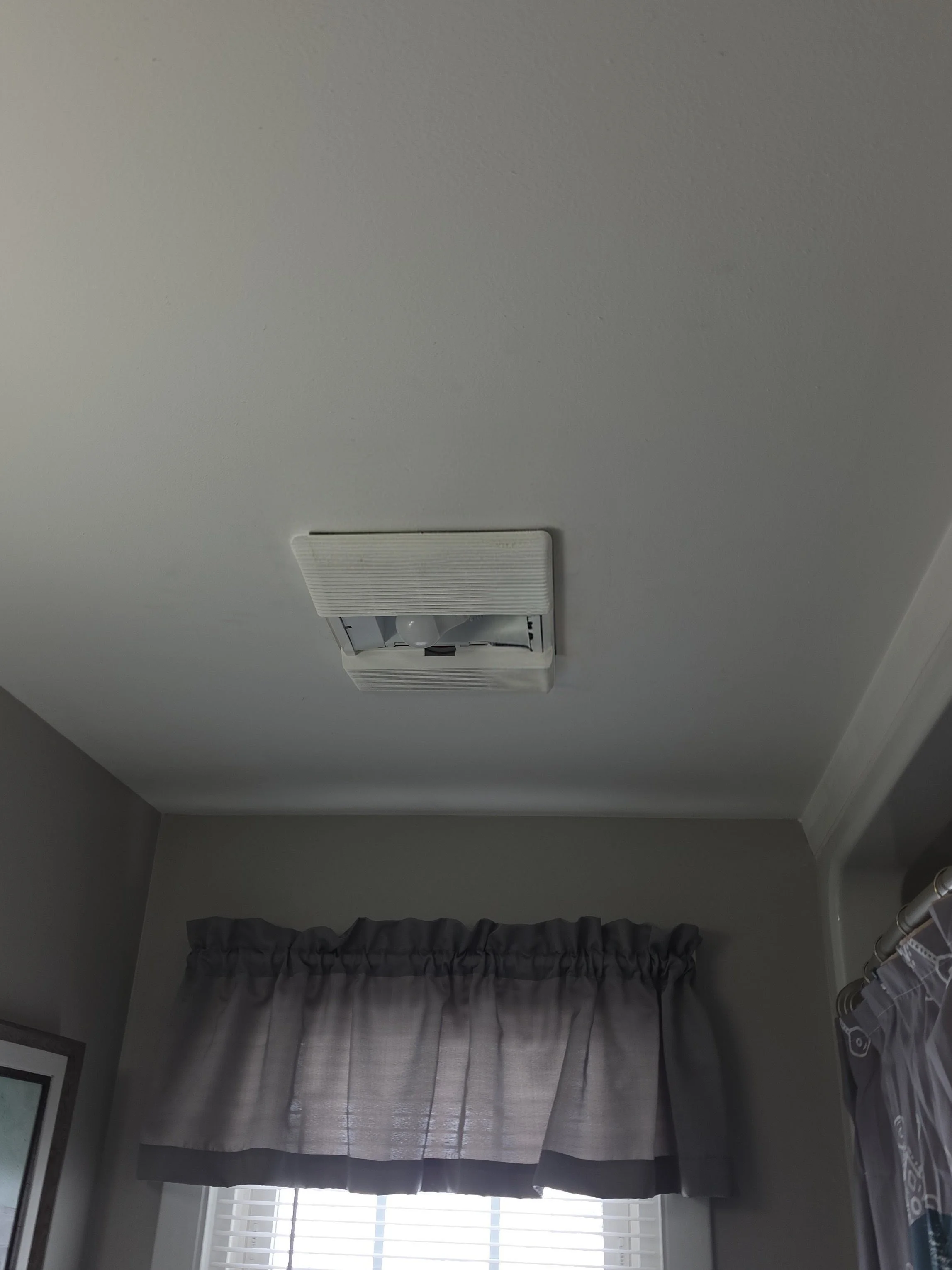 Ceiling vent fan with partial view of window and gray curtains in a room.
