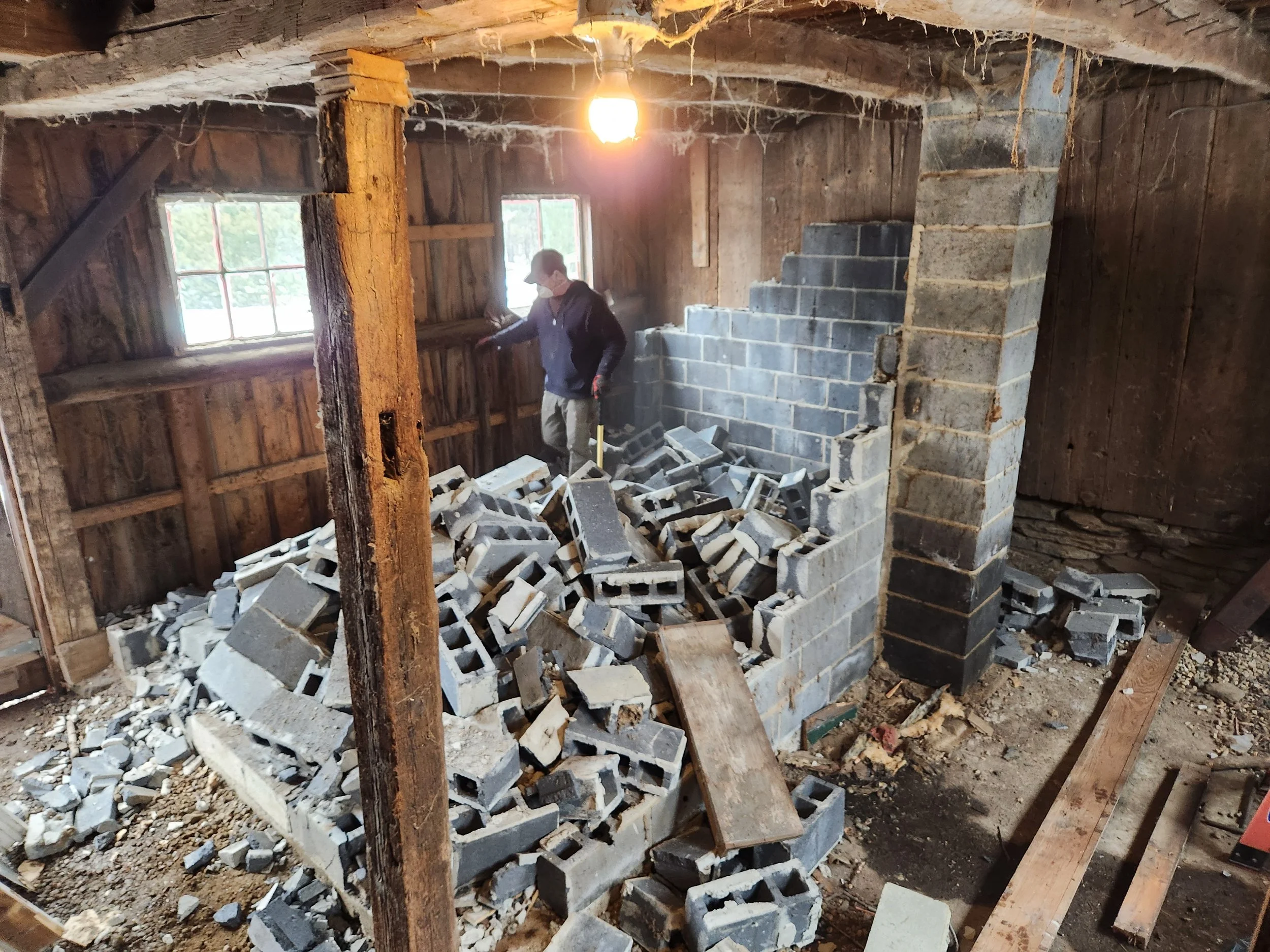 A person constructing a brick wall inside a rustic, unfinished wooden barn with exposed beams and walls. The floor is cluttered with loose bricks and construction debris, and a single light bulb hangs from the ceiling.
