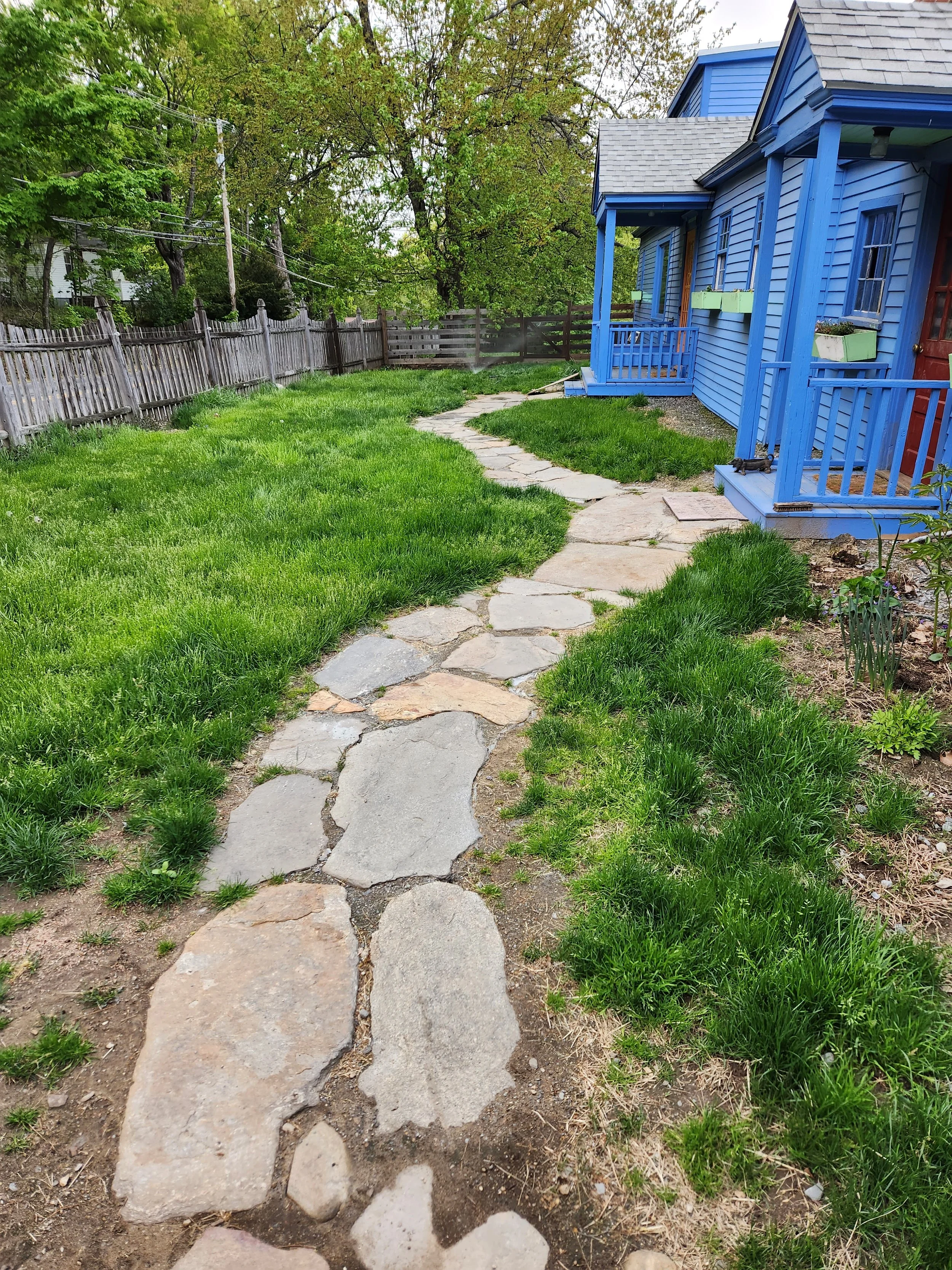 A stone pathway winding through a backyard with green grass, next to a blue house with porch steps, surrounded by a wooden fence and trees.