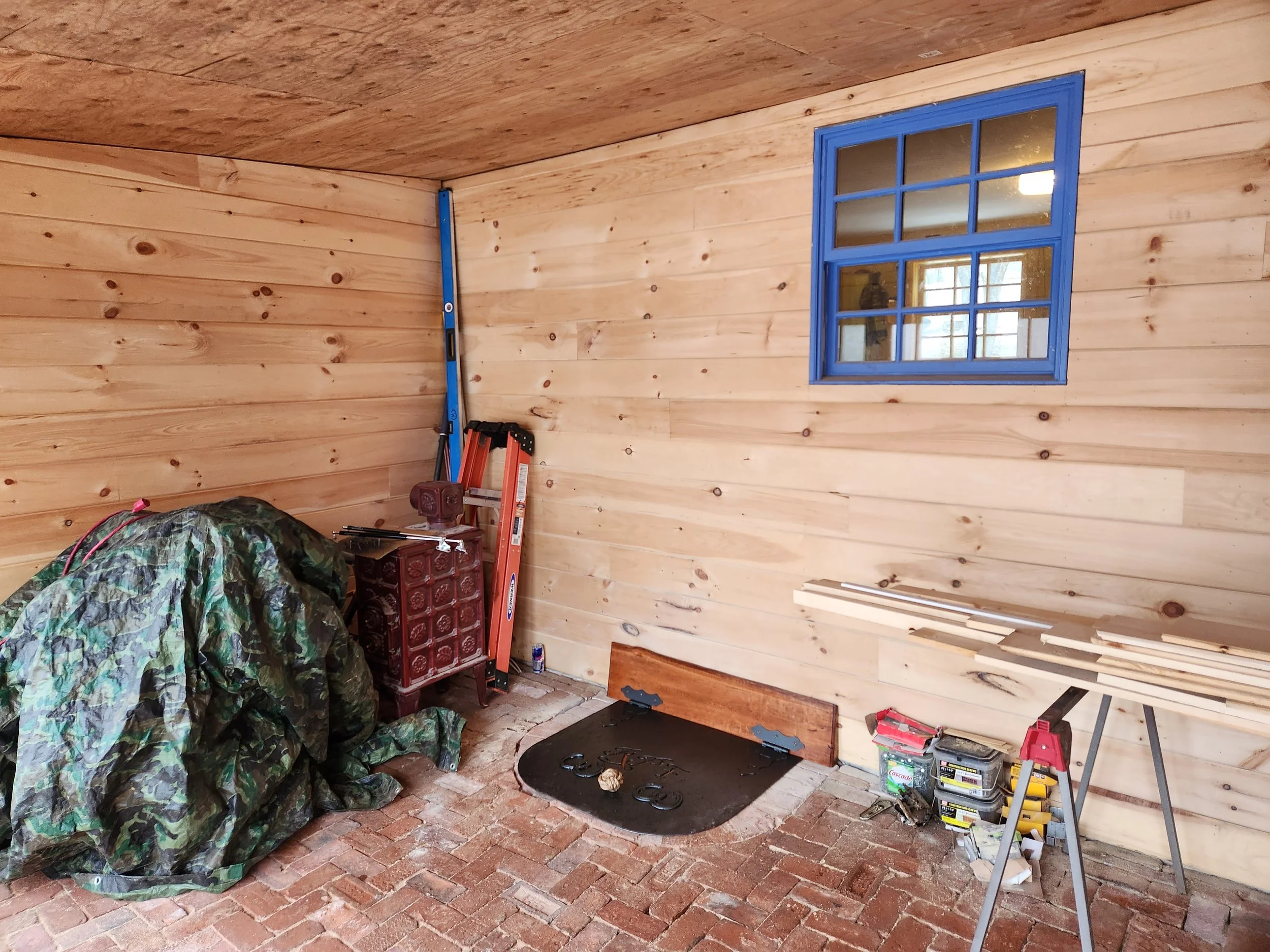 An unfinished room with wooden walls and ceiling, a small window with blue trim, a work table with wood planks, a sawhorse, and tools, bricks on the floor, and a covered object.