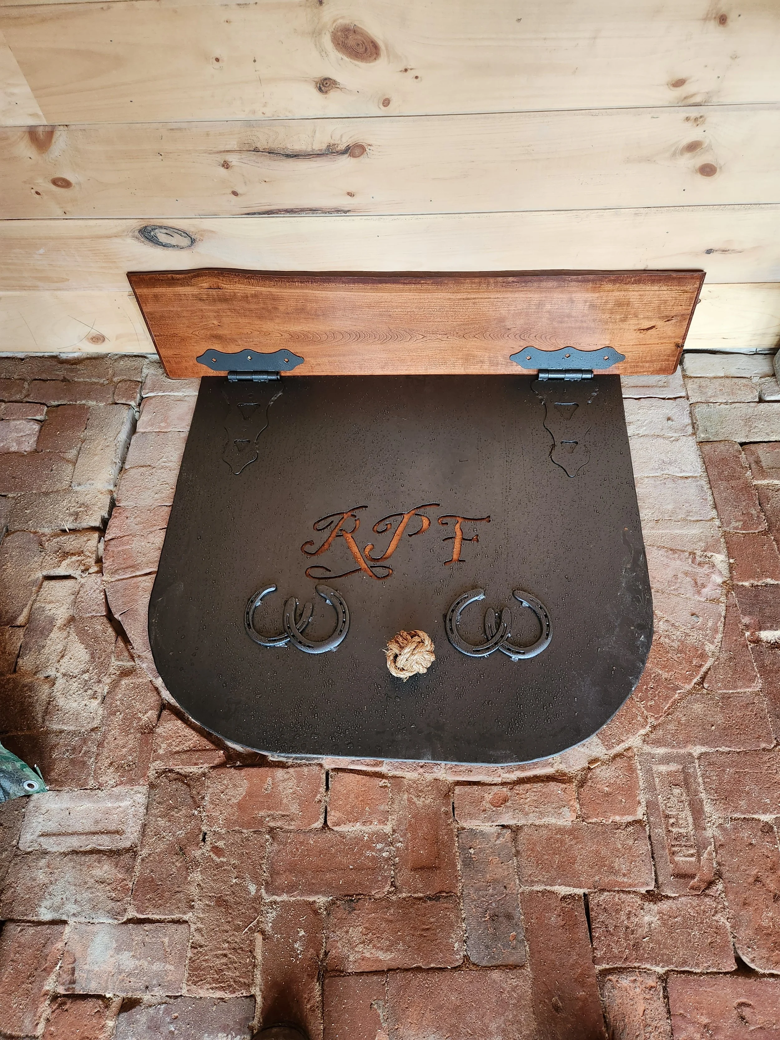A black metal horseshoe-shaped door mat with the initials 'KPF' carved in the center, placed on a brick floor, with a wooden door on top and mounted with hinges.