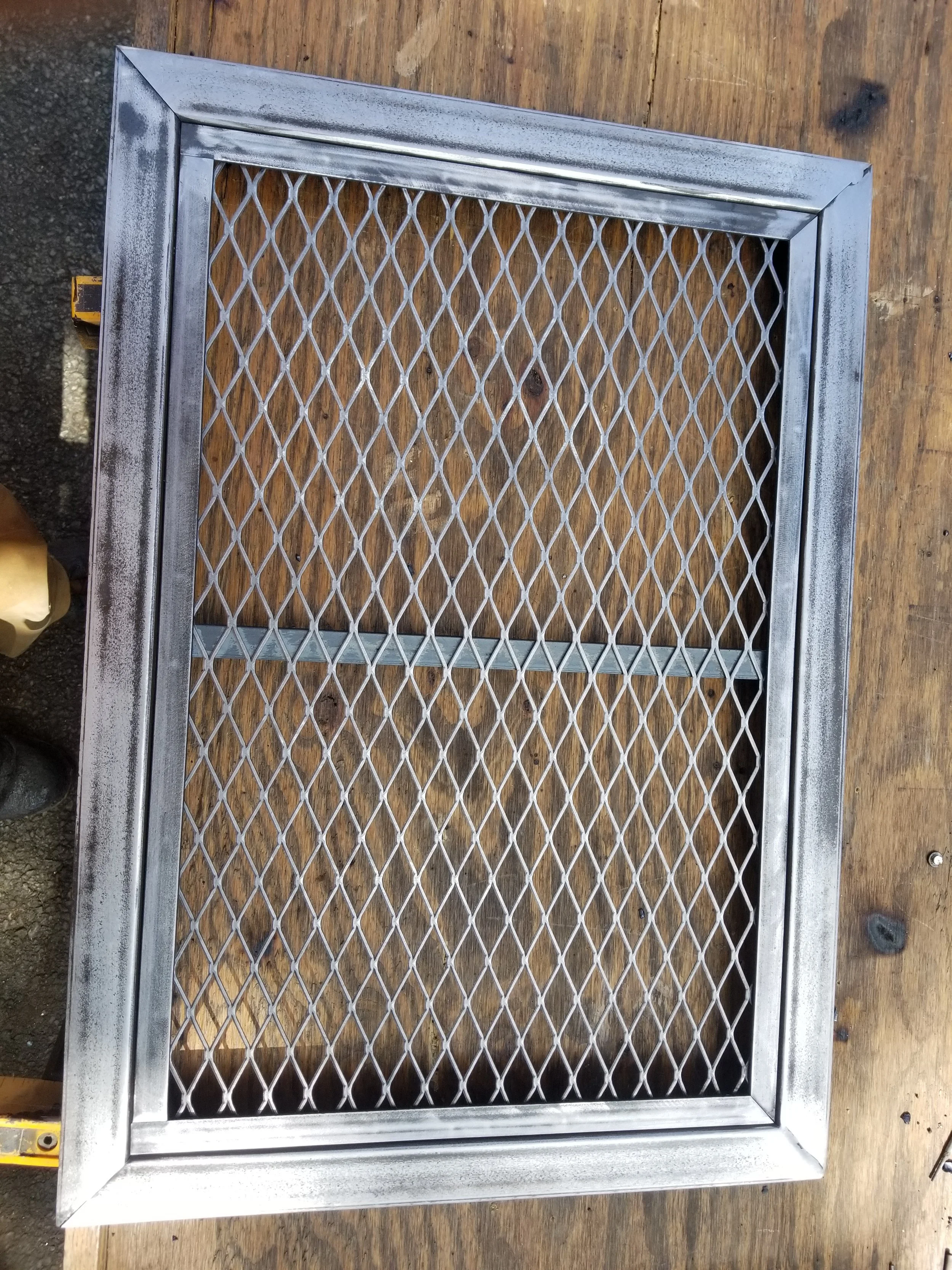 A metal catwalk grate with a diamond pattern, framed with metal, laying on a wooden surface.