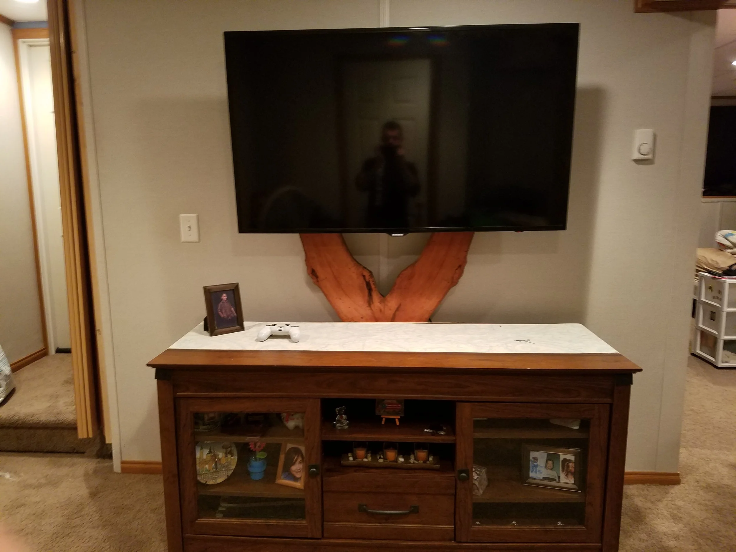 Living room entertainment setup with a mounted flat-screen TV above a wooden console, which has glass doors displaying framed photographs, decorations, and small items. A framed photo and a white gaming controller are on top of the console.