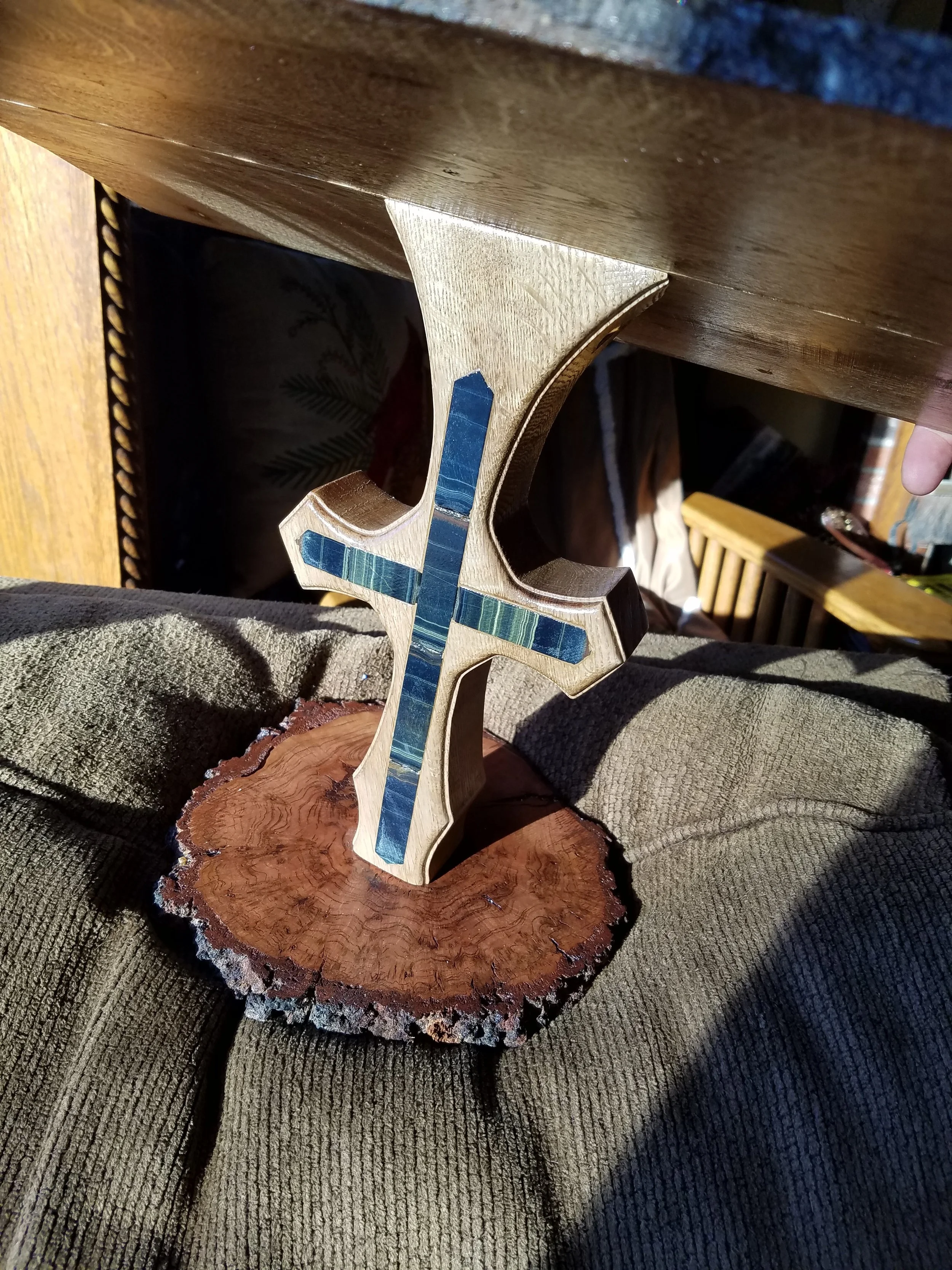A wooden cross with inlay details, mounted on a polished wood slice, placed on a gray fabric surface with a wooden table nearby.