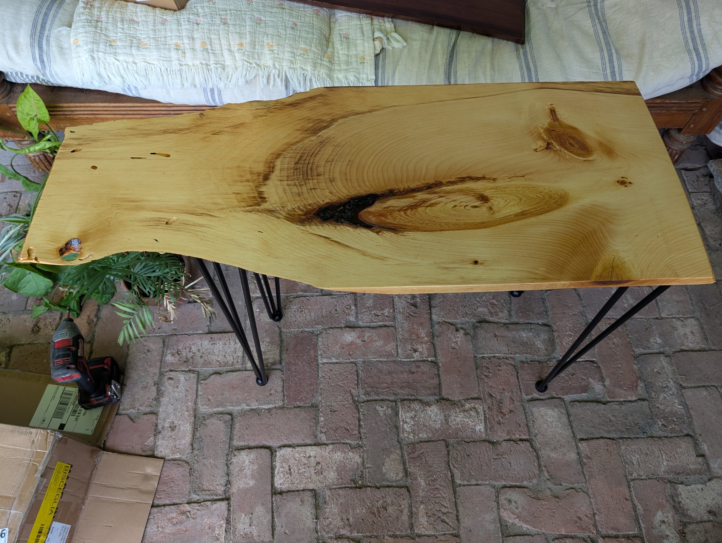 A long wooden table with a natural, unfinished look supported by black metal hairpin legs, placed on a brick patio.