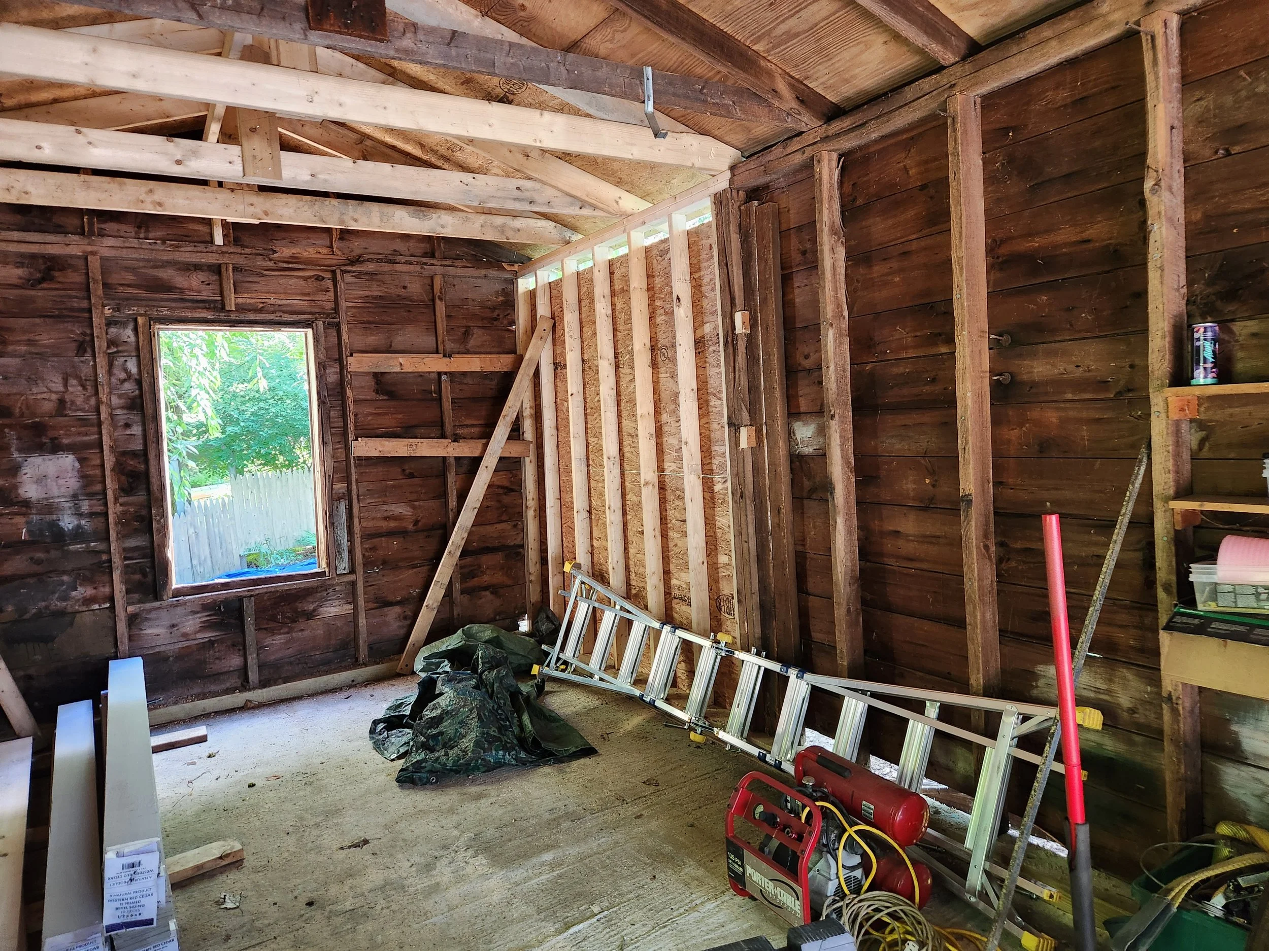 Interior of a house under renovation showing unfinished walls with exposed wooden framing, a ladder, and construction tools on the floor. A window reveals a green outdoor area.