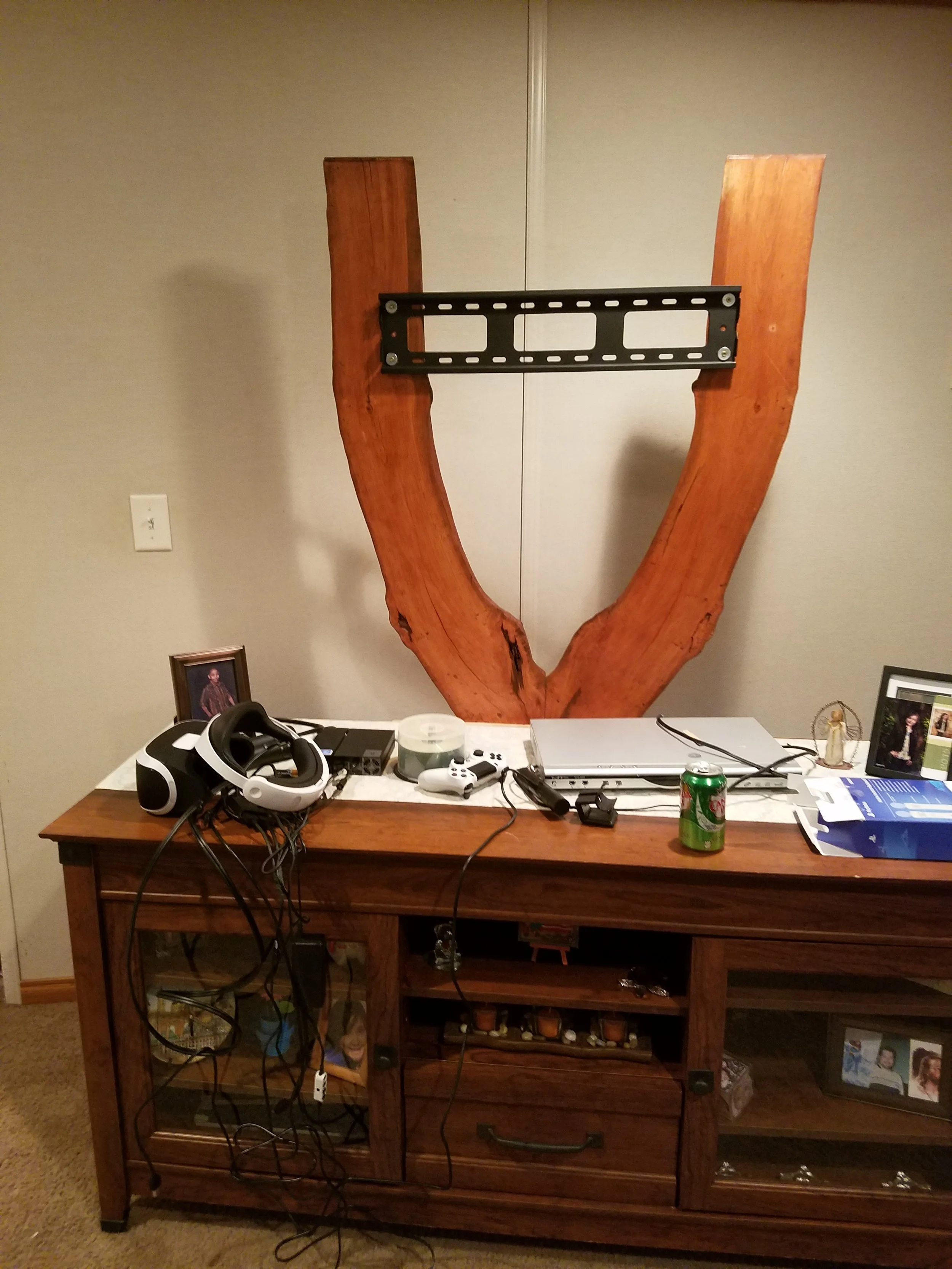 Photograph of a living room wall with a large wooden 'U' shape structure mounted above a wooden sideboard. The sideboard has various electronic devices, a seltzer can, framed photographs, and clutter including wires and a headset.