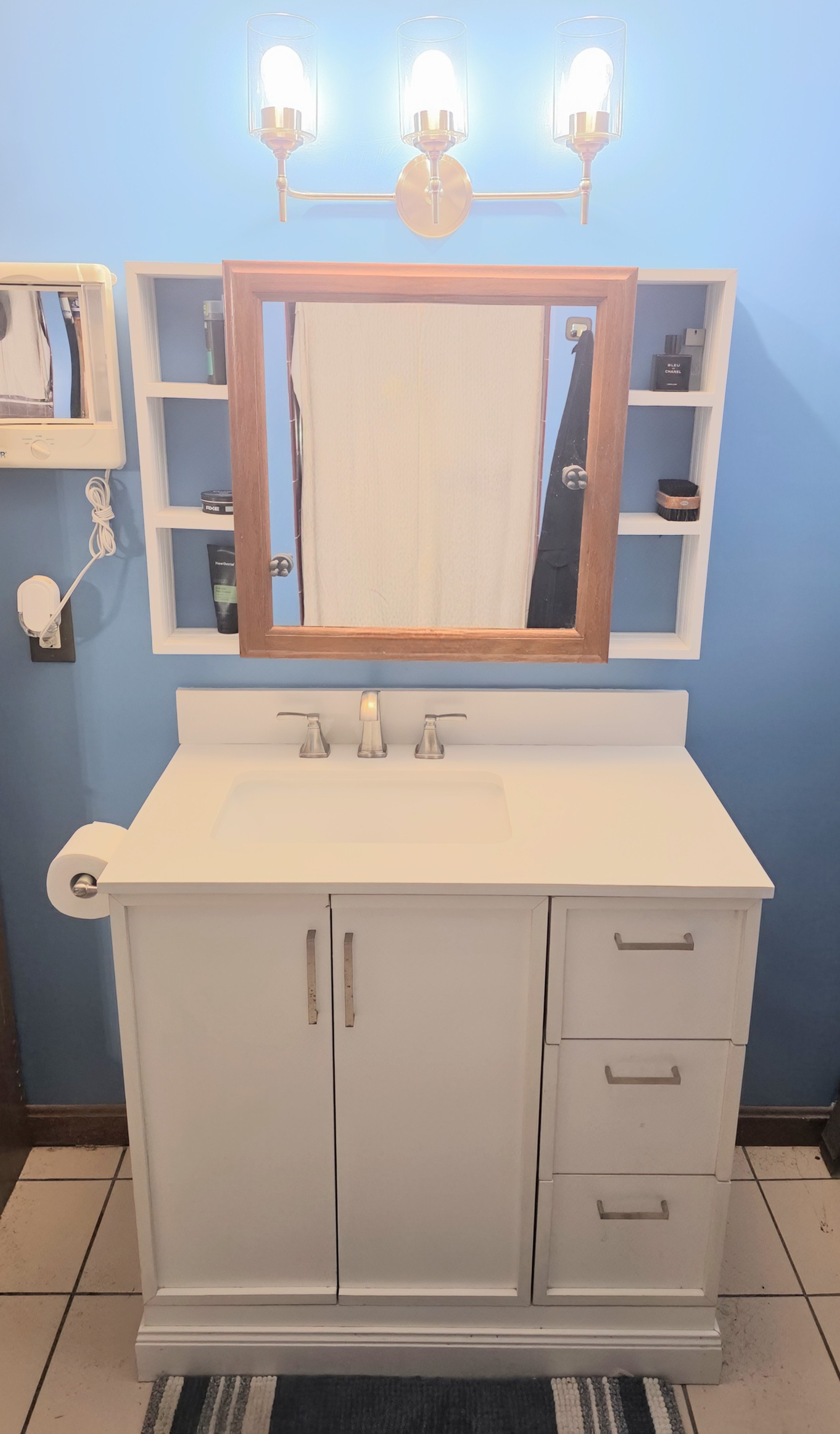 Bathroom vanity with a white cabinet, a rectangular mirror with a wooden frame, and a three-light fixture above. Blue wall, shelves with toiletries, and a mirror on the wall.