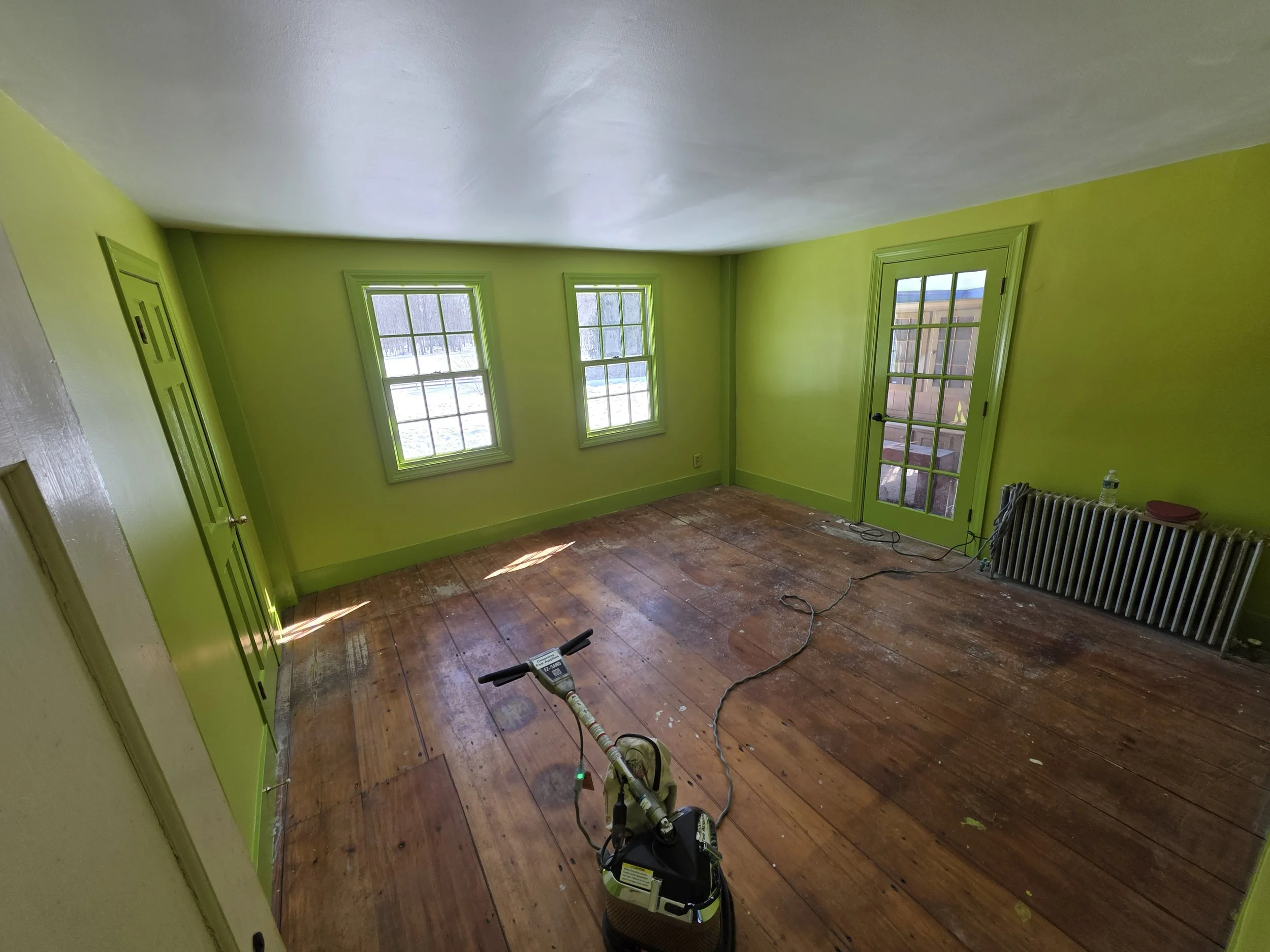 Empty room with bright lime green walls, three windows, and a glass door leading outside; wooden floor with some marks and equipment, including a steamer and cables.