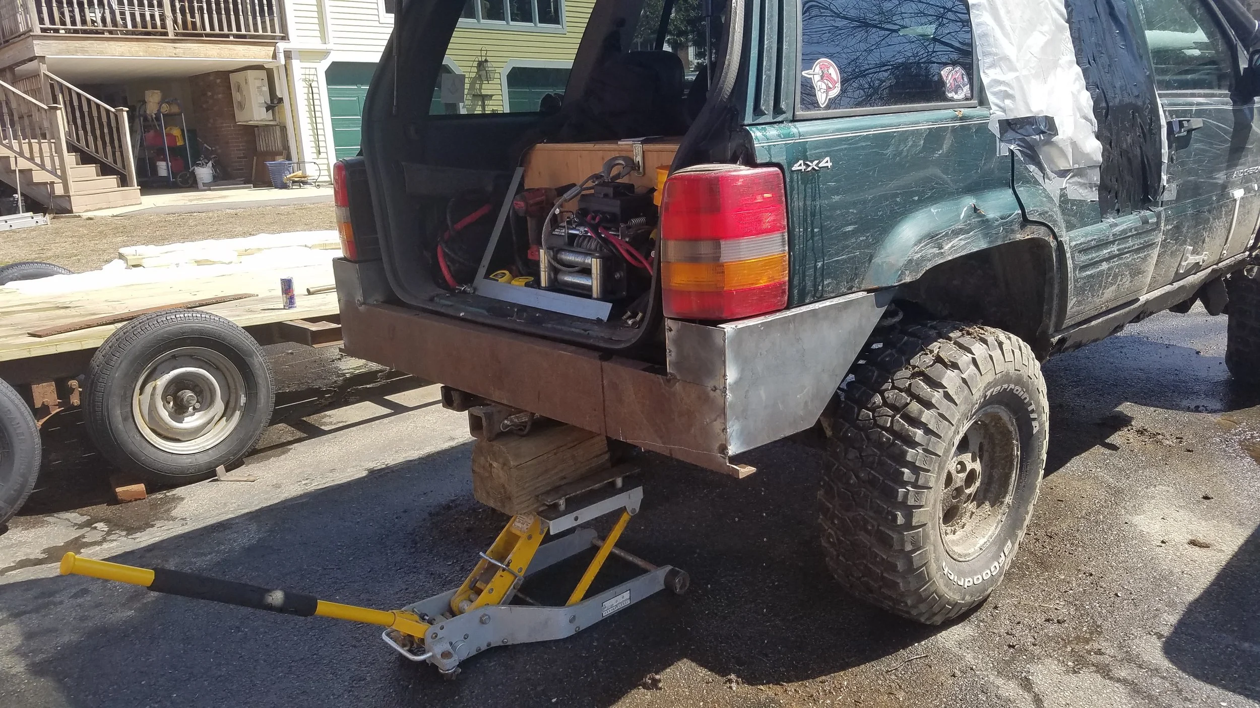 A green off-road vehicle with damaged side panels and an open rear hatch, showing some mechanical equipment inside. The vehicle is raised on a hydraulic jack and placed on a wooden block, with a flatbed trailer attached nearby in a parking lot with residential buildings in the background.