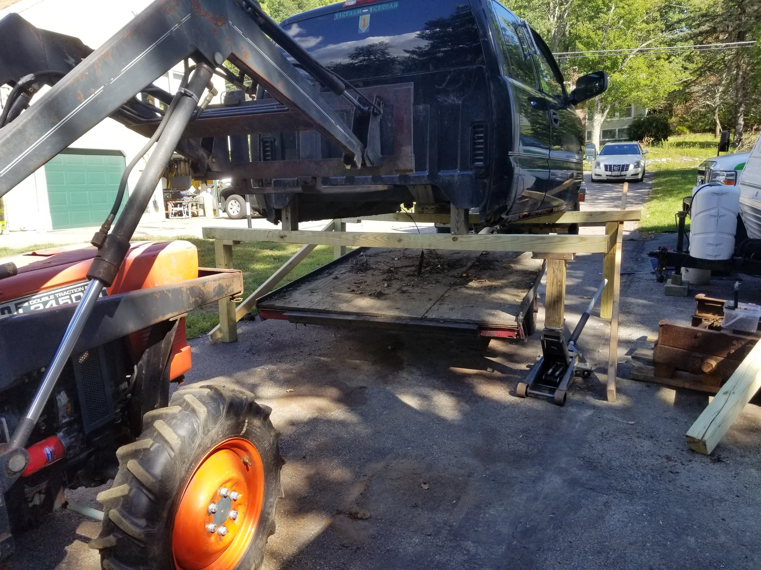 A black truck is lifted on a wooden frame in a driveway, with a tractor nearby and other vehicles parked in the background.