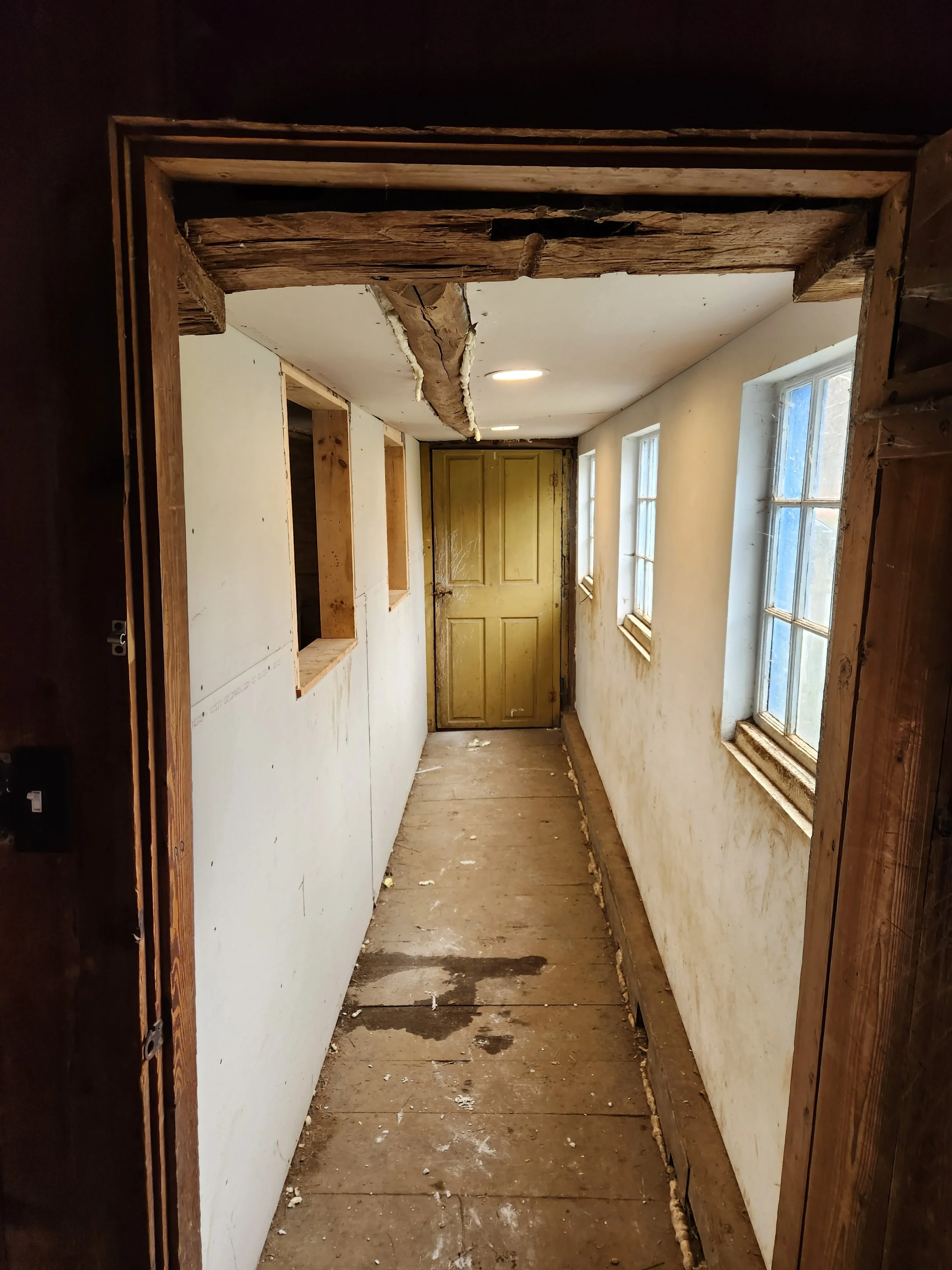 A narrow hallway under renovation with exposed wooden framing and a damaged ceiling, featuring three small windows on the right wall and a closed yellow door at the end.