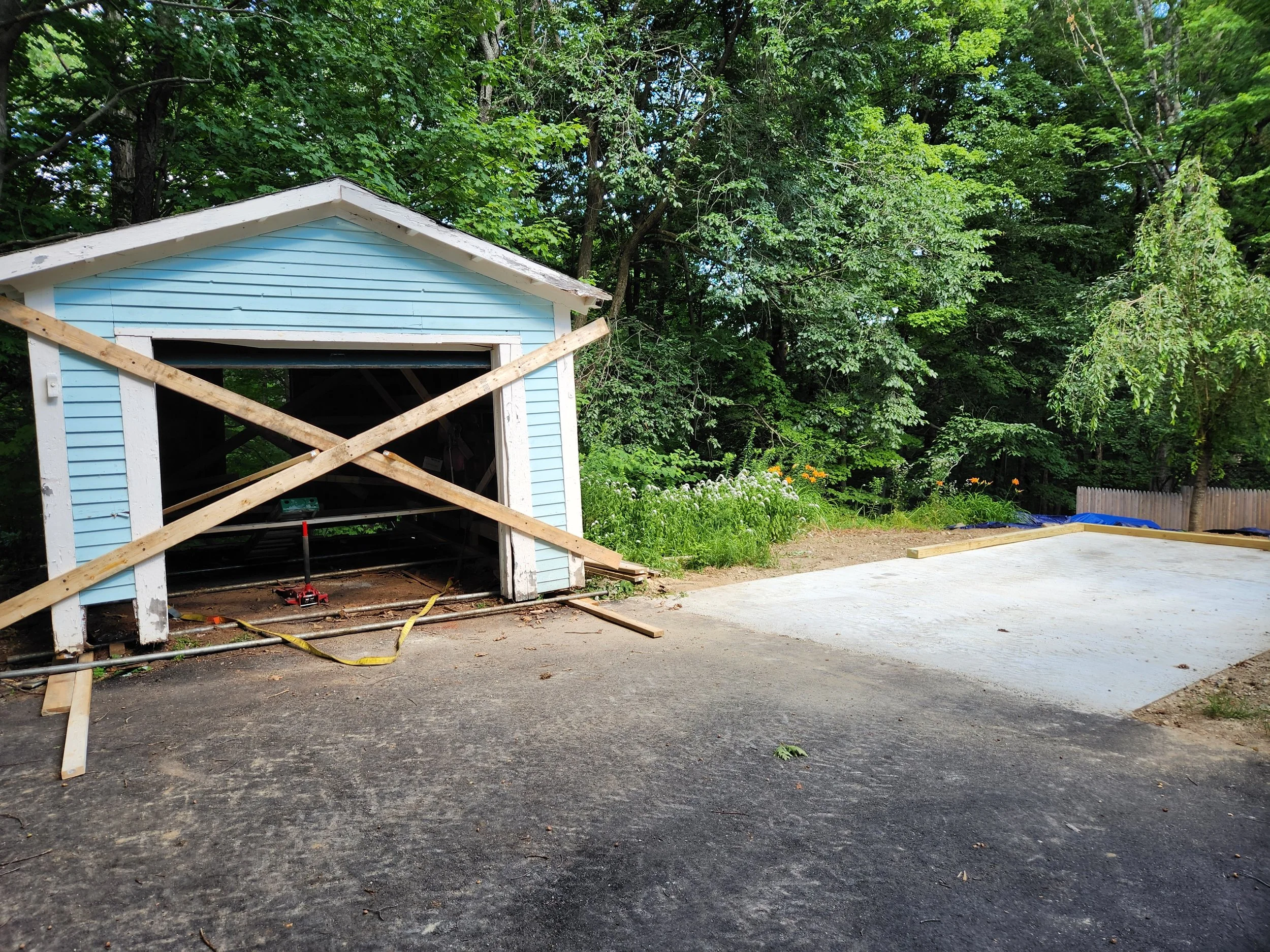A small blue shed with a white trim is under renovation, with wooden braces across the front. Next to it is a concrete slab, likely for a new driveway or patio, with a wooded area in the background.