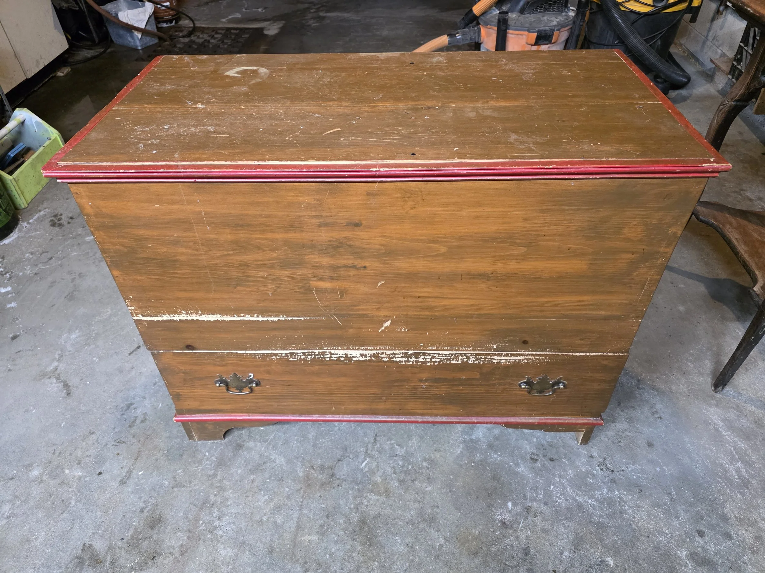 A vintage wooden dresser with two drawers, worn surface, and red trim on the edges, placed on a concrete floor in a garage or workshop setting.