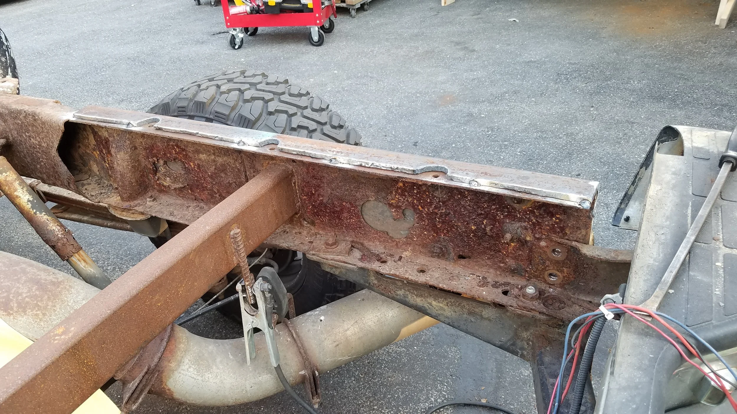 Rusty vehicle frame with disconnected wiring, mounted on a workbench with a tire in the background.