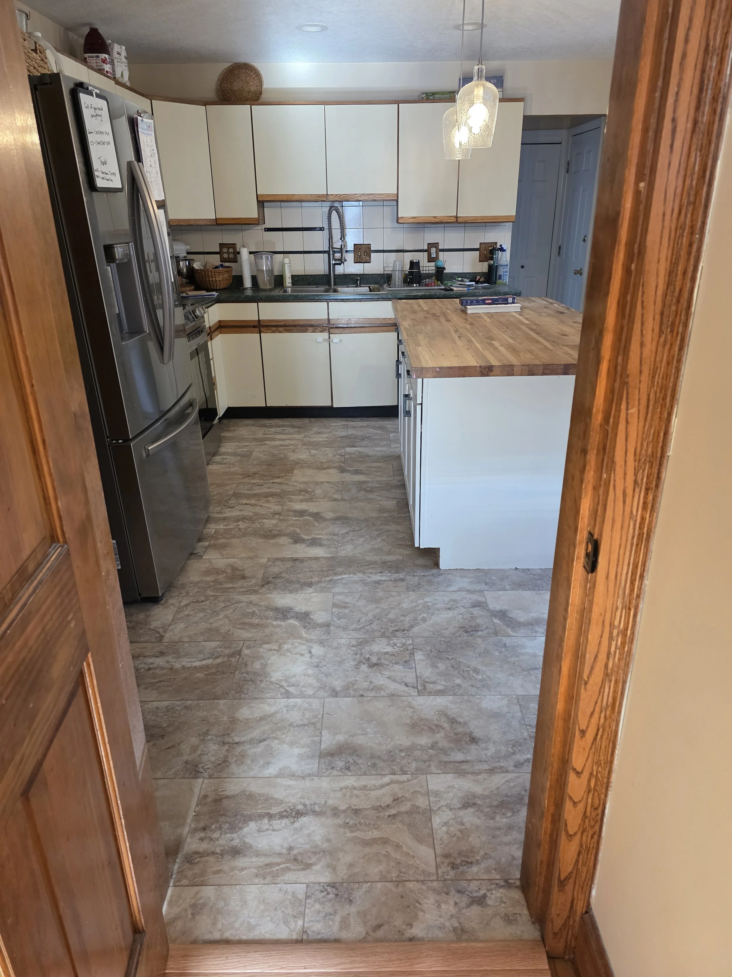 View of a kitchen with white cabinets, a stainless steel refrigerator, a wooden kitchen island, tiled floor, and a pendant light hanging from the ceiling.